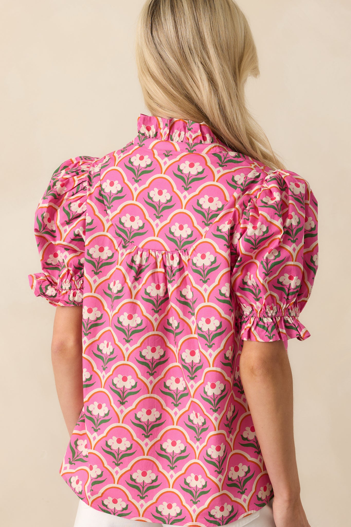 Up-close photo of model showing the back of a pink top that features a pink multicolor geometric floral print, ruffle notch v-neckline, functional button front, short puff sleeves, and ruffle elastic cuffs.
