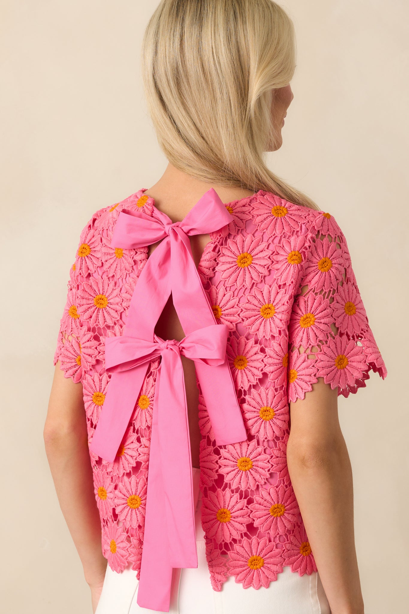Up-close photo of modeling wearing and showing the back of a pink and orange floral embroidered lace overlay, hot pink lining, daisy petal scalloped hems, open self-tie bow back with two bows.