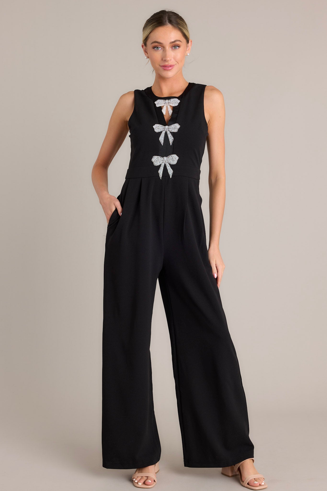Full body view of this black jumpsuit that features a round neckline, a discrete back zipper, three ornate bows embellished with faux ivory pearls and rhinestones, a chest cutout, a thick waistband, subtle pleats, functional hip pockets, and a wide leg design.