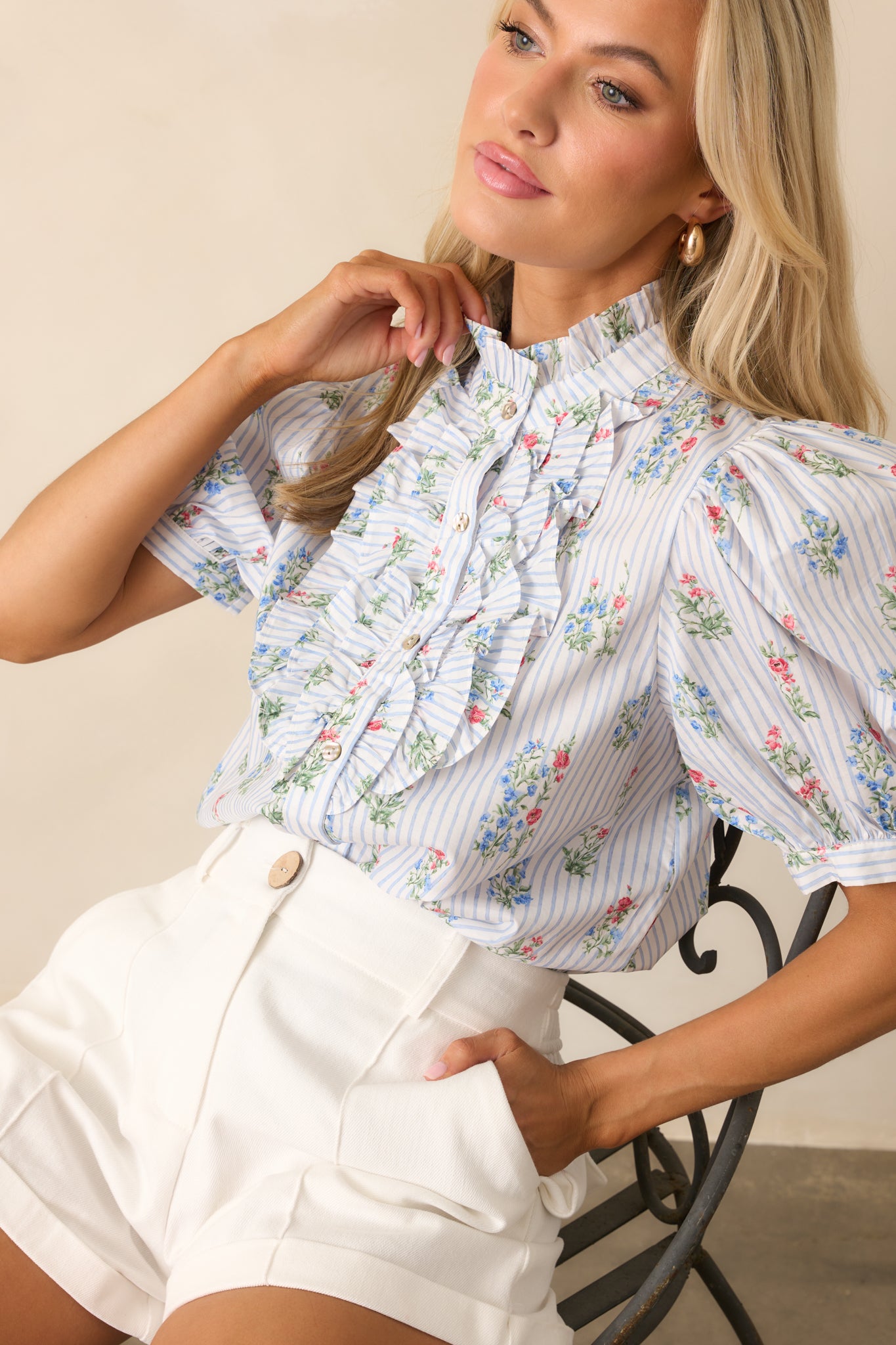 Close-up of woman adjusting the high ruffled neckline of a light blue striped blouse with floral print and front ruffle detail, wearing white high-waisted shorts.