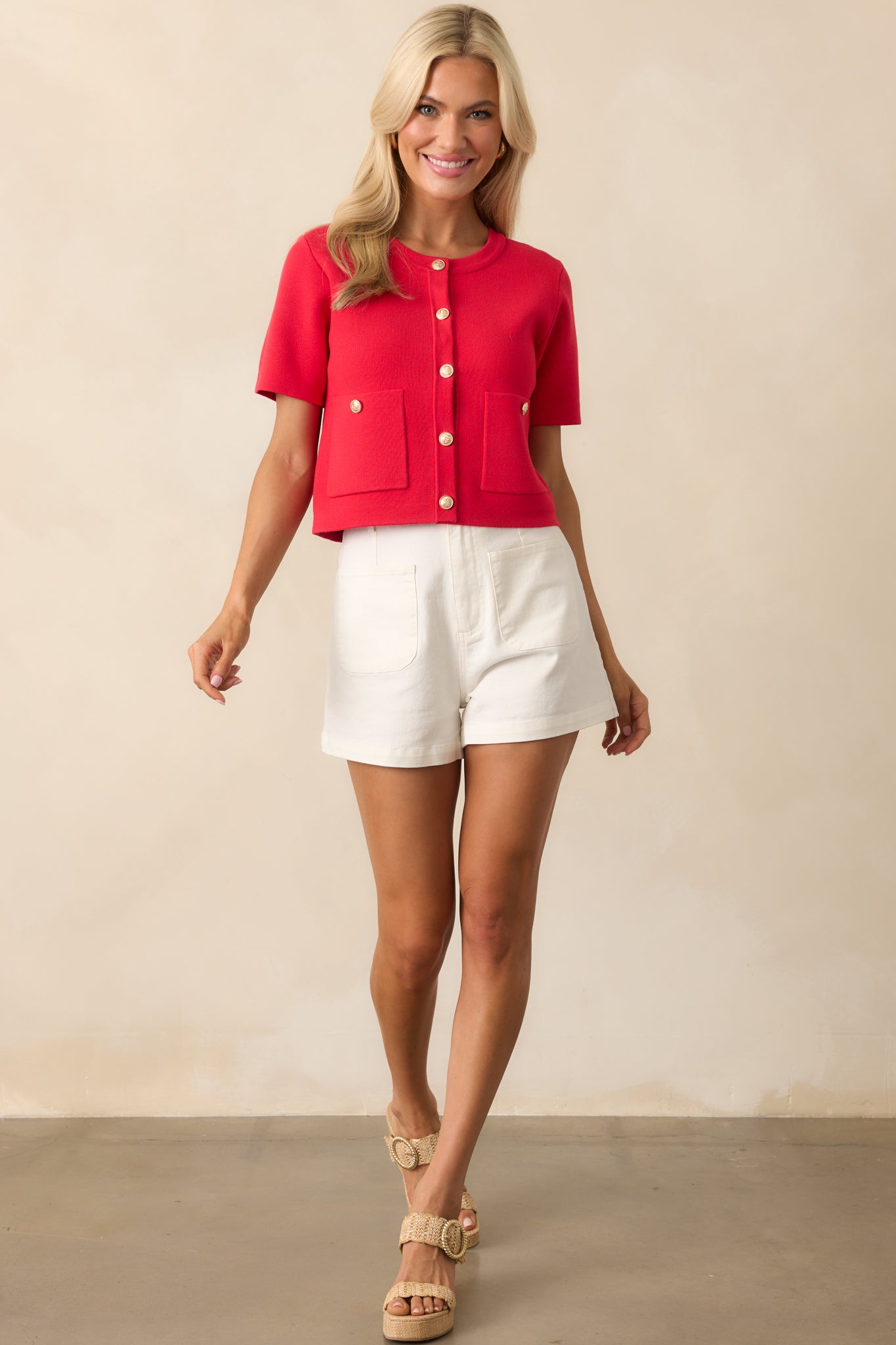Woman standing and smiling in a bright red cropped cardigan with gold buttons, white high-waisted shorts, and woven platform sandals.