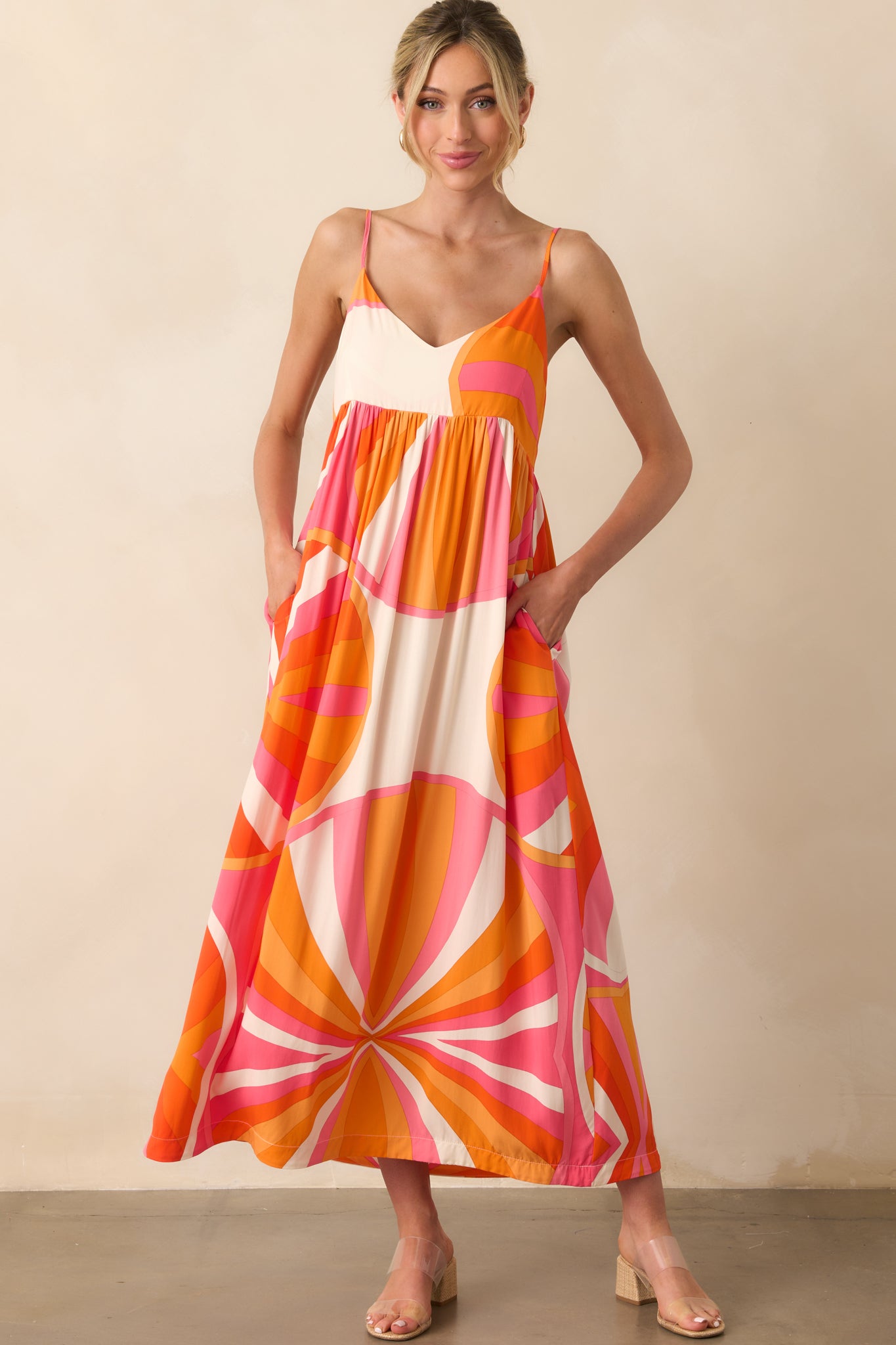 Full body photo of model wearing maxi dress that features an orange and pink abstract print, lightweight 100% rayon fabric, v-neckline, adjustable spaghetti straps, relaxed fit, and functional pockets.