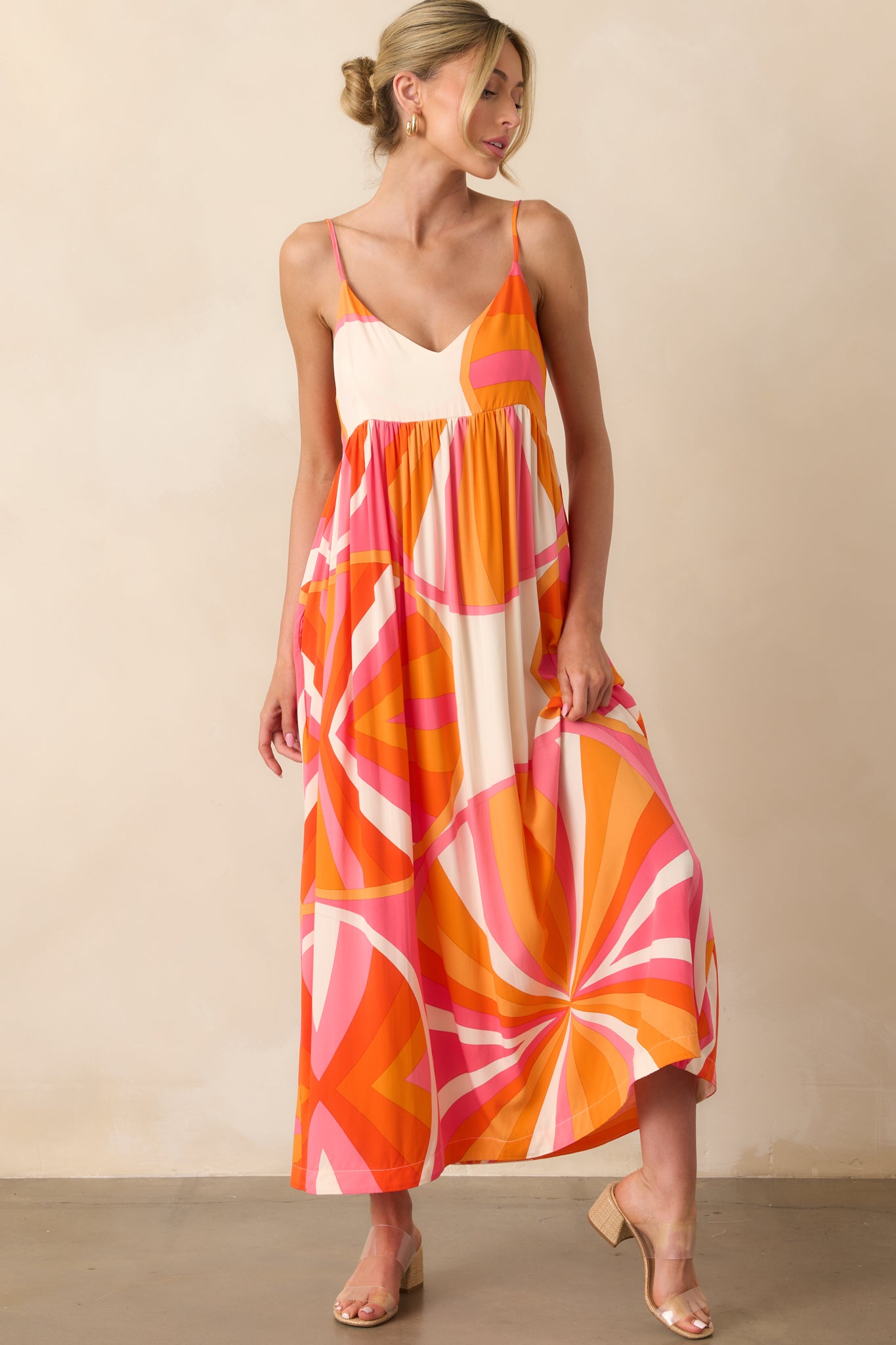 Full body photo of model wearing maxi dress that features an orange and pink abstract print, lightweight 100% rayon fabric, v-neckline, adjustable spaghetti straps, relaxed fit, and functional pockets.