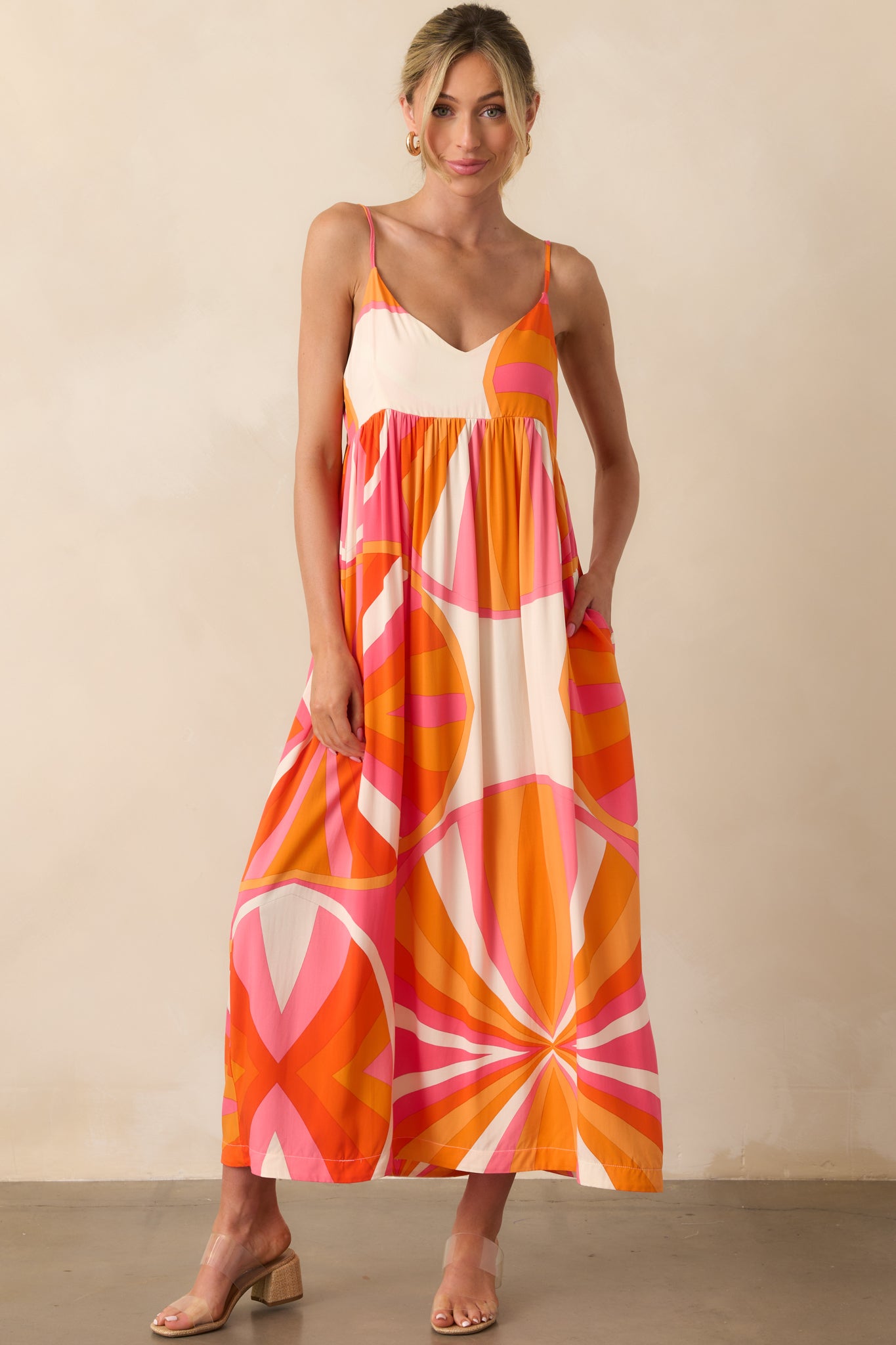 Full body photo of model wearing maxi dress that features an orange and pink abstract print, lightweight 100% rayon fabric, v-neckline, adjustable spaghetti straps, relaxed fit, and functional pockets.