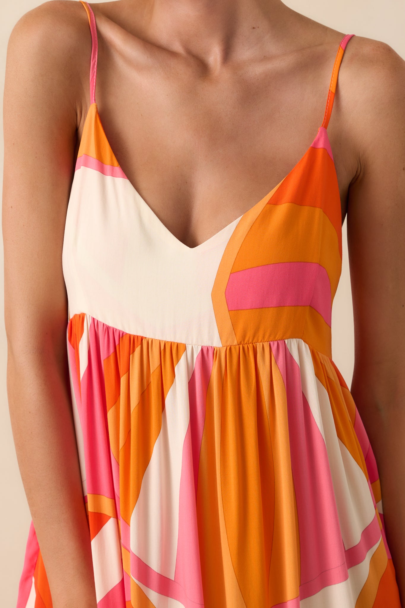 Up-close photo of model wearing maxi dress that features an orange and pink abstract print, v-neckline, adjustable spaghetti straps, relaxed fit, and functional pockets.