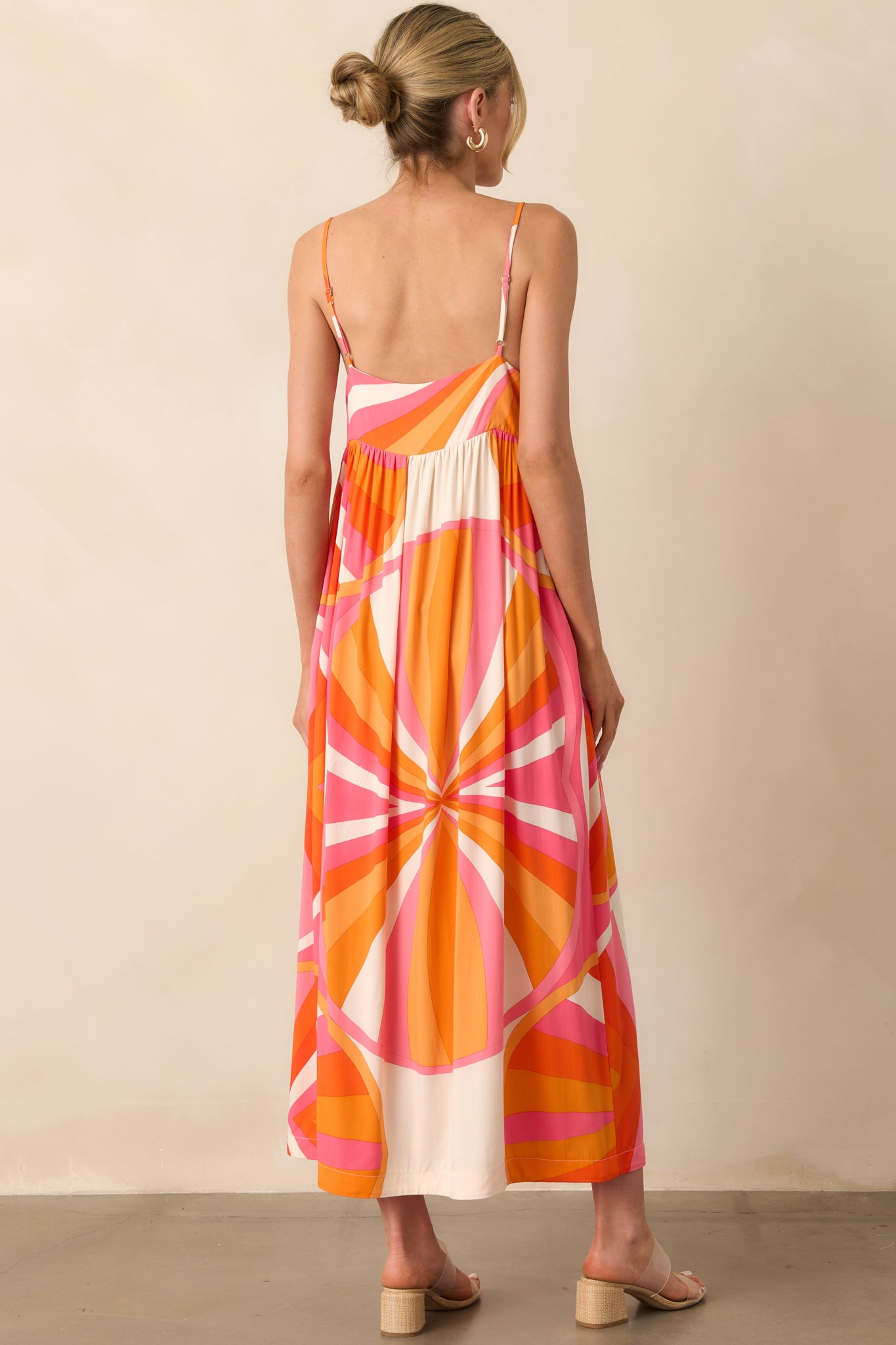 Full body photo of model showing the back of a maxi dress that features an orange and pink abstract print, lightweight 100% rayon fabric, v-neckline, adjustable spaghetti straps, relaxed fit, and functional pockets.