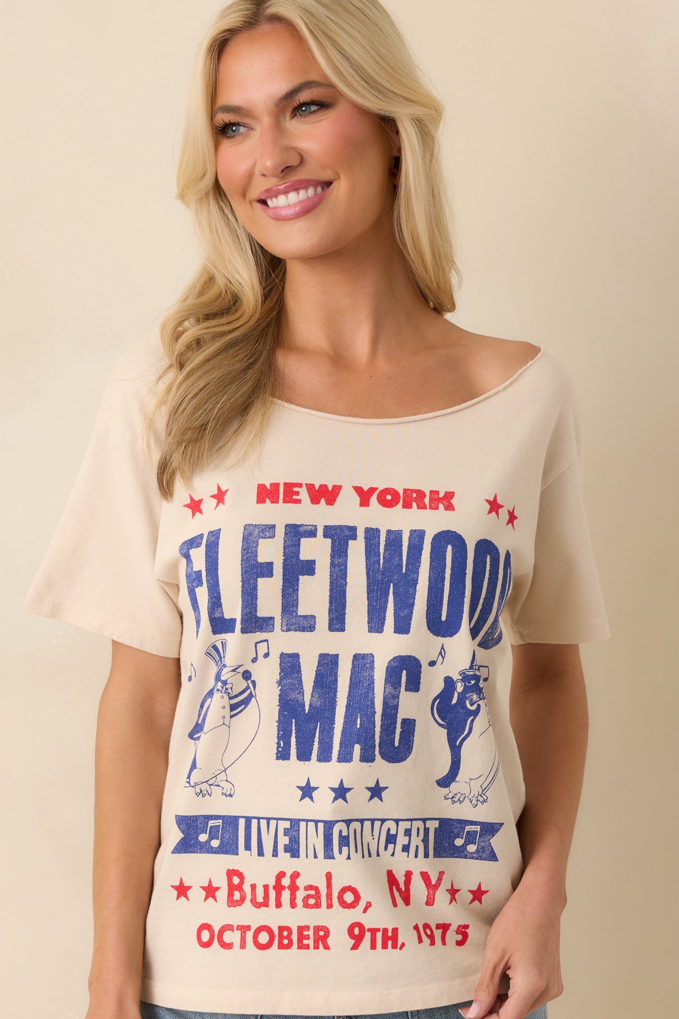 Up close photo of model wearing a beige tee that features a graphic of a Fleetwood Mac 1975 New York show poster with penguin mascots, an off-the-shoulder neckline, short sleeves, and a relaxed fit.