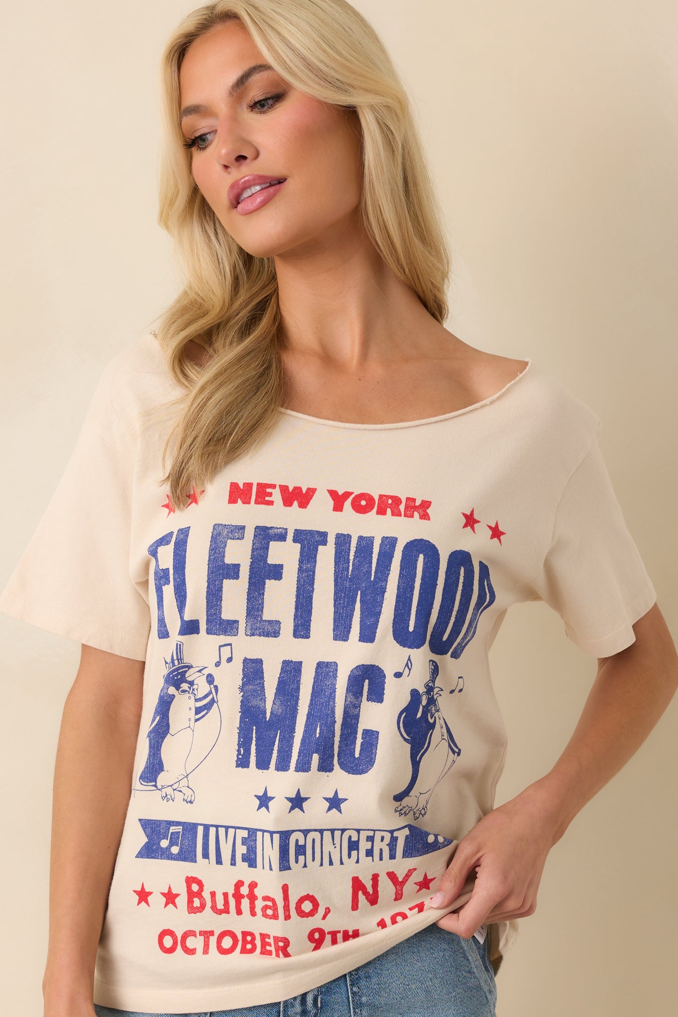 Up close photo of model wearing a beige tee that features a graphic of a Fleetwood Mac 1975 New York show poster with penguin mascots, an off-the-shoulder neckline, short sleeves, and a relaxed fit.