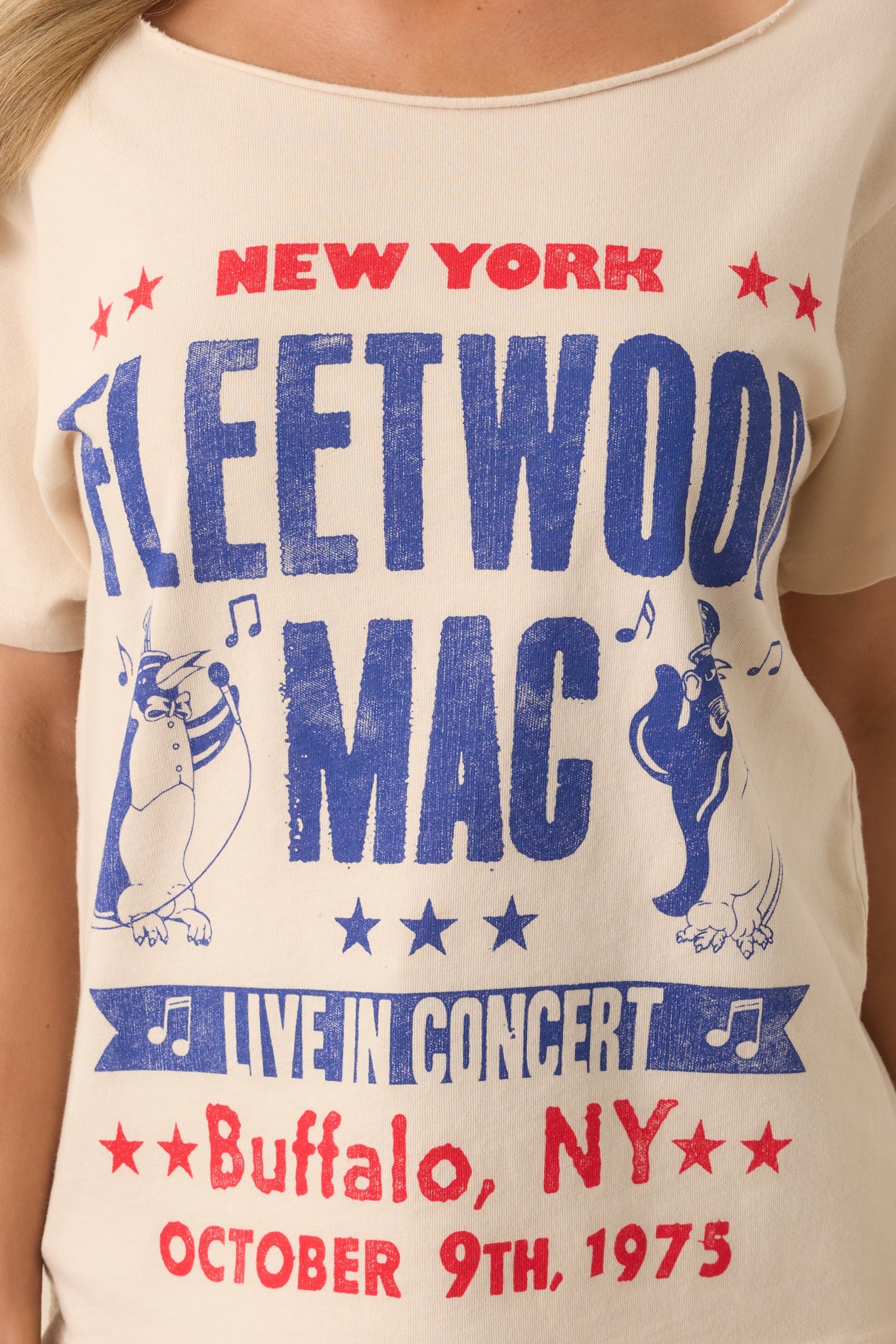 Up close photo a beige tee that features a graphic of a Fleetwood Mac 1975 New York show poster with penguin mascots, an off-the-shoulder neckline, short sleeves, and a relaxed fit.