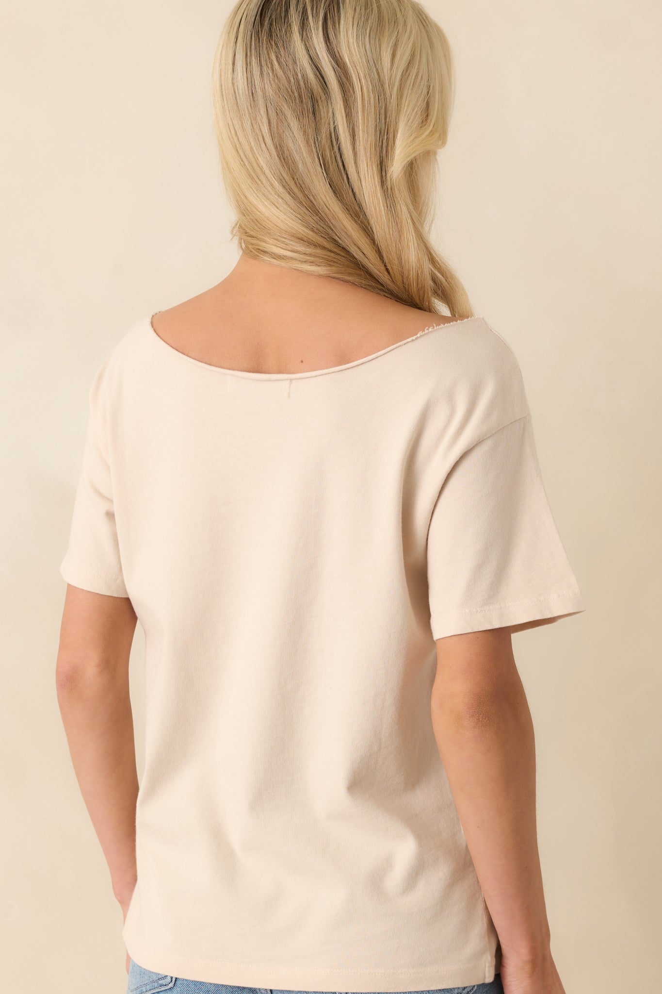 Up close photo of back of model wearing a beige tee that features a graphic of a Fleetwood Mac 1975 New York show poster with penguin mascots on the front, an off-the-shoulder neckline, short sleeves, and a relaxed fit.