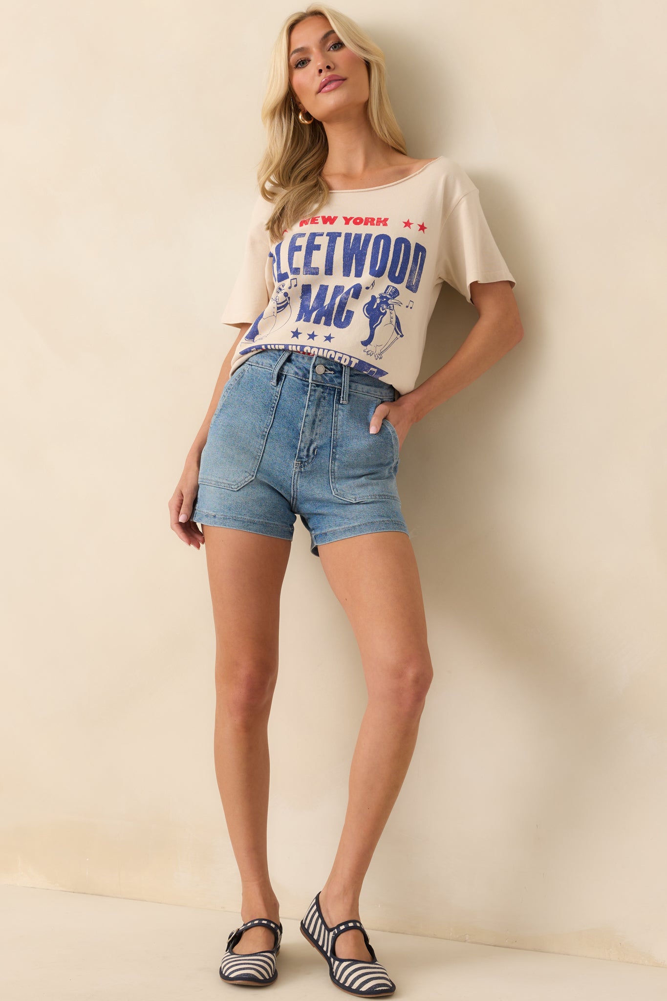 A full body photo of model wearing a beige tee that features a graphic of a Fleetwood Mac 1975 New York show poster with penguin mascots, and denim shorts.