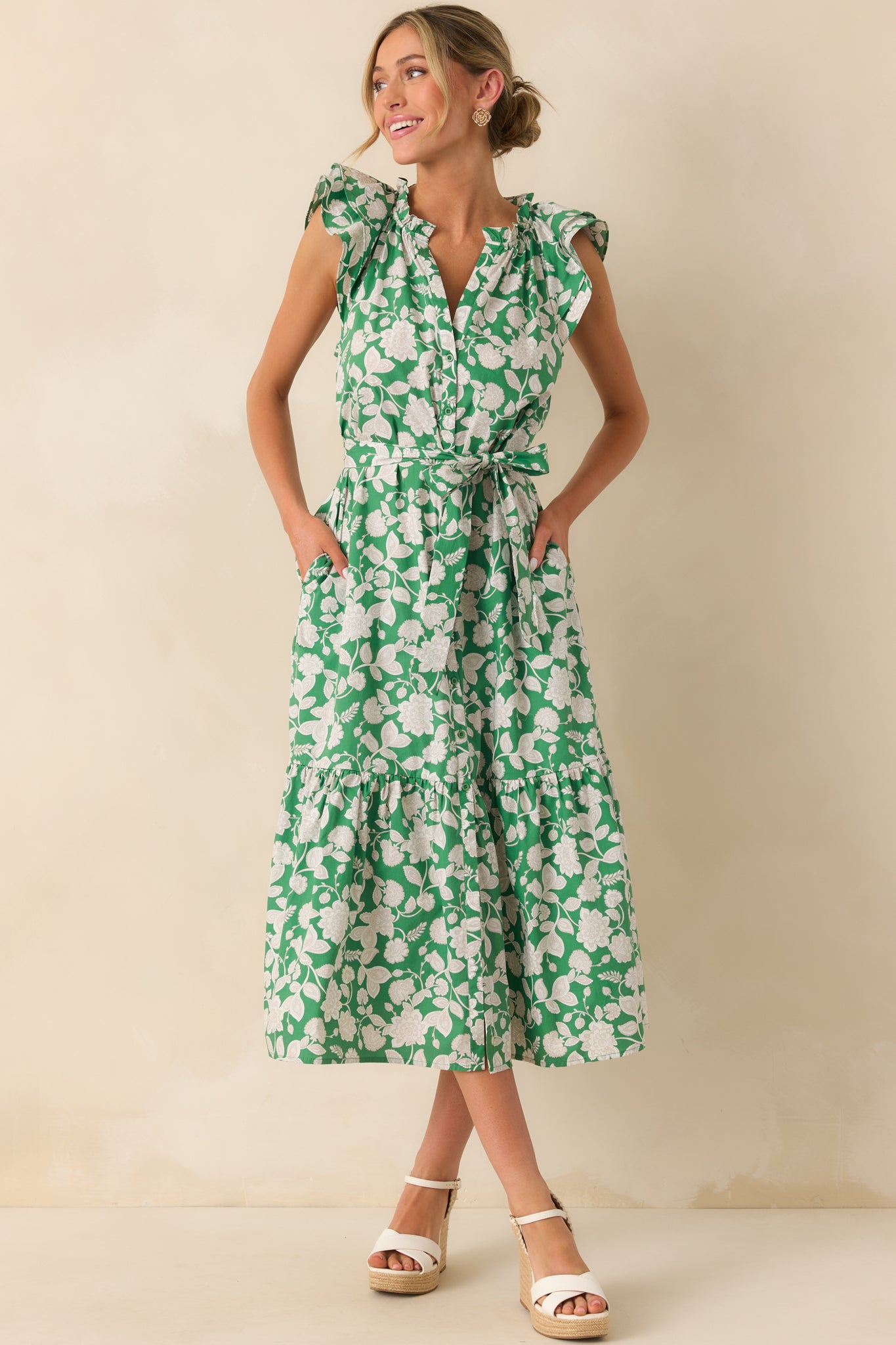 Model standing and wearing a green maxi dress that features a ruffle notch v-neckline, layered flutter sleeves, functional button front, optional self-tie belt, 100% cotton fabric, bottom tiered skirt, and functional pockets.