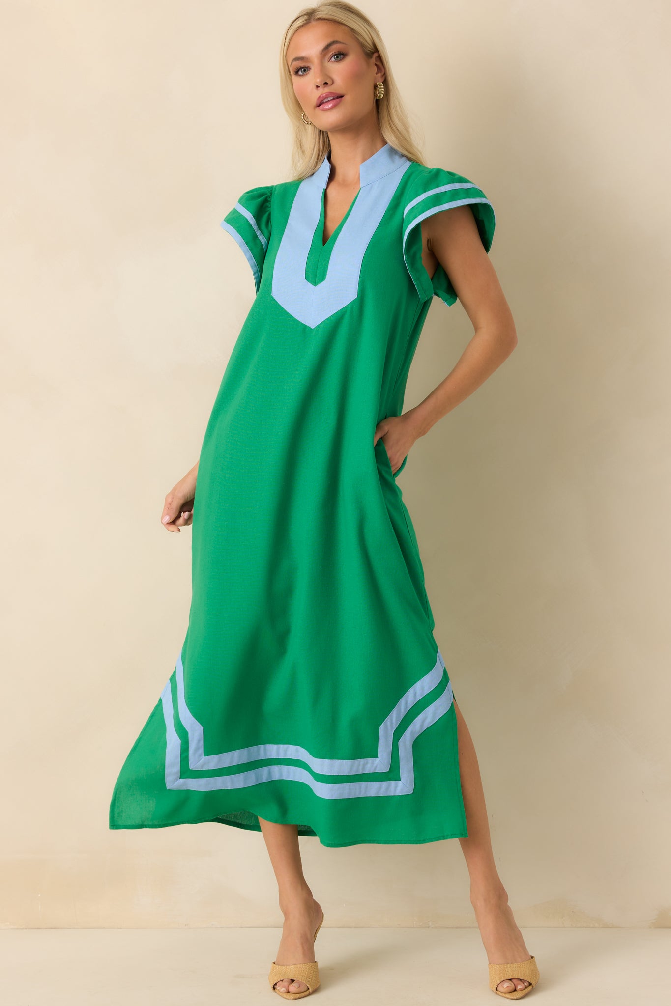 Full body photo of model wearing a green maxi dress that features a light blue contrast trim throughout, notch v-neckline, short flutter sleeves, and bottom hem vent slits.