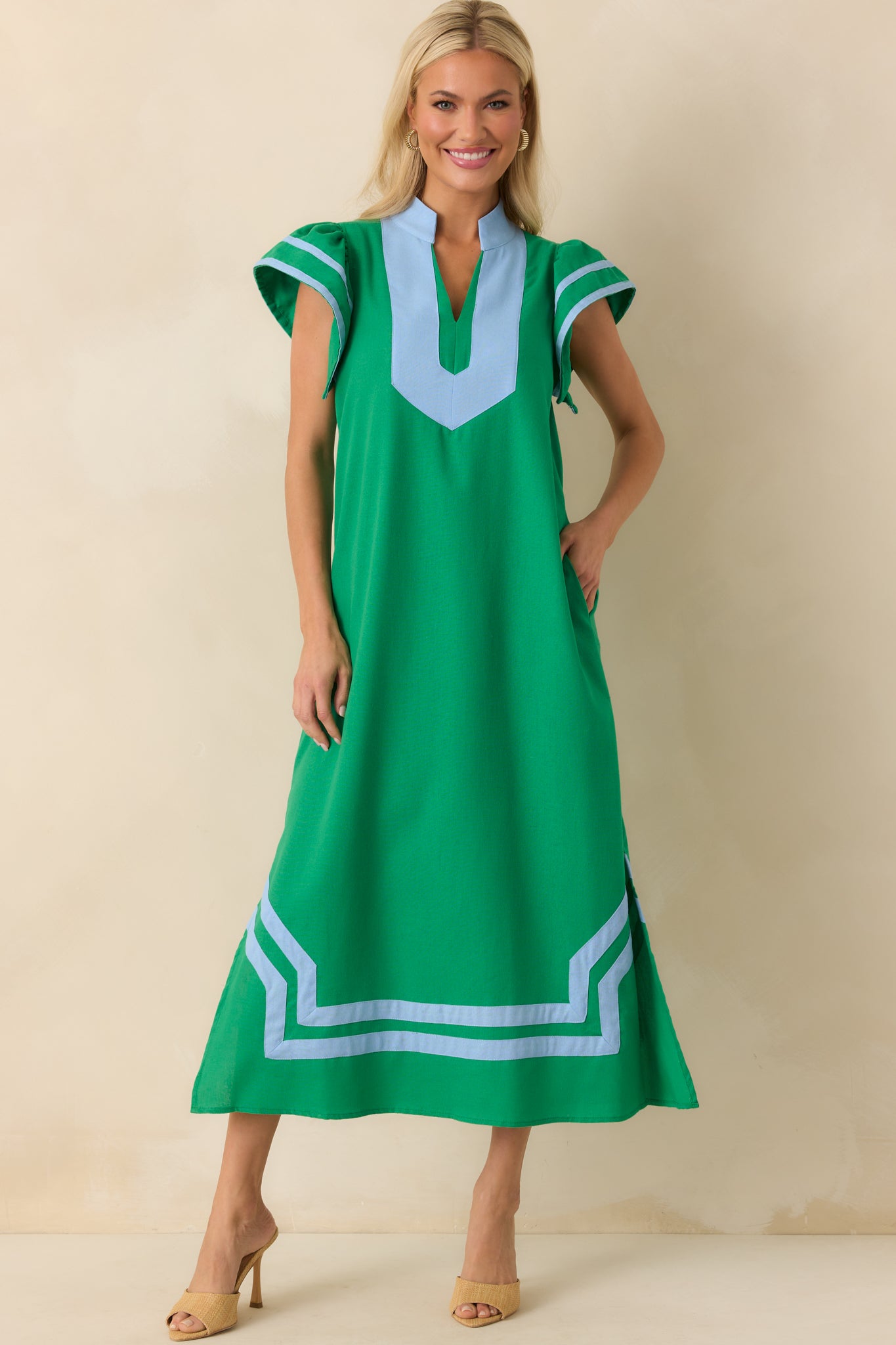Full body photo of model wearing a green maxi dress that features a light blue contrast trim throughout, notch v-neckline, short flutter sleeves, and bottom hem vent slits.