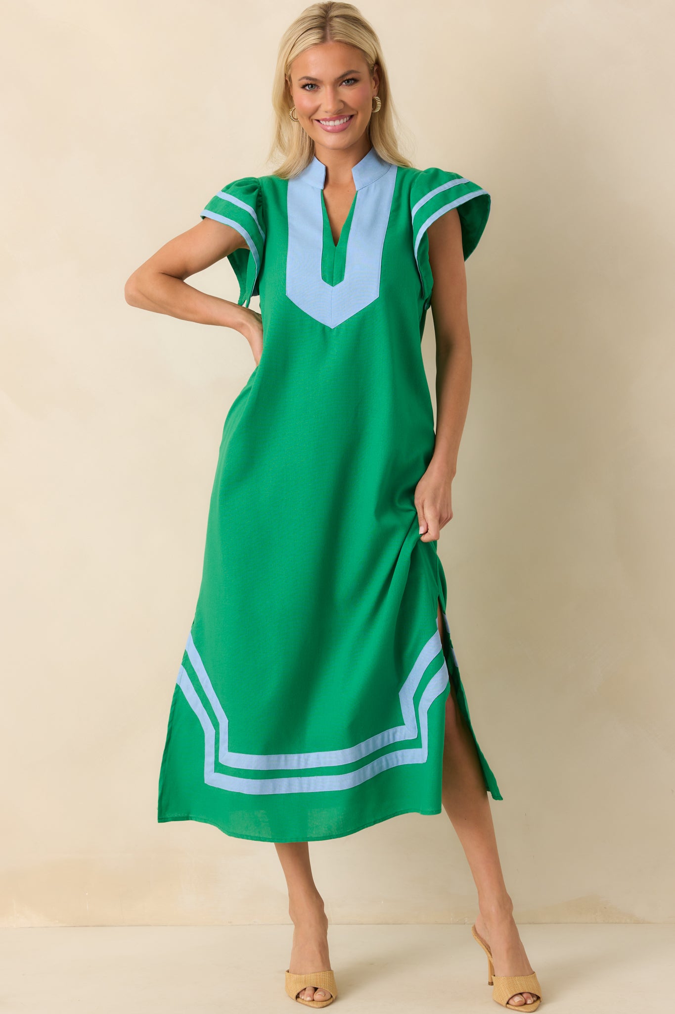Full body photo of model with hand on her hip wearing a green maxi dress that features a light blue contrast trim throughout, notch v-neckline, short flutter sleeves, and bottom hem vent slits.