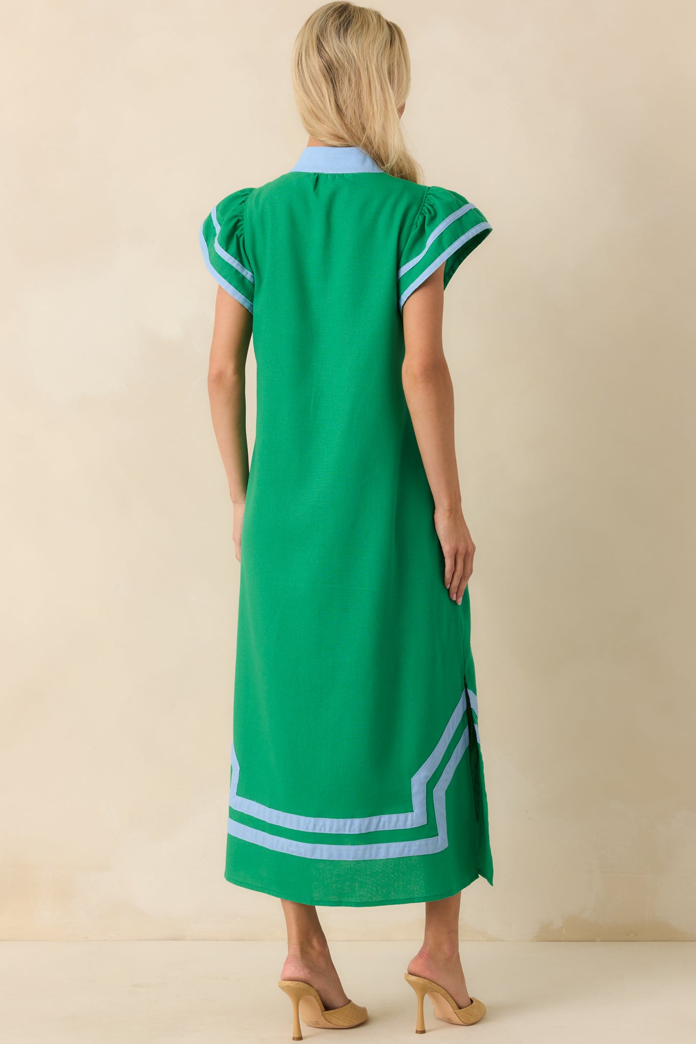 Full body photo of the back of a model wearing a green maxi dress that features a light blue contrast trim throughout, notch v-neckline, short flutter sleeves, and bottom hem vent slits.