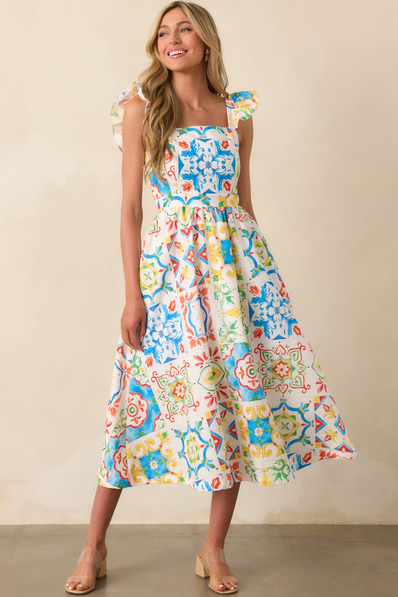 A blue midi dress featuring flutter sleeves and a squared neckline, shown in full front view with the Sicilian tile design across the entire dress.