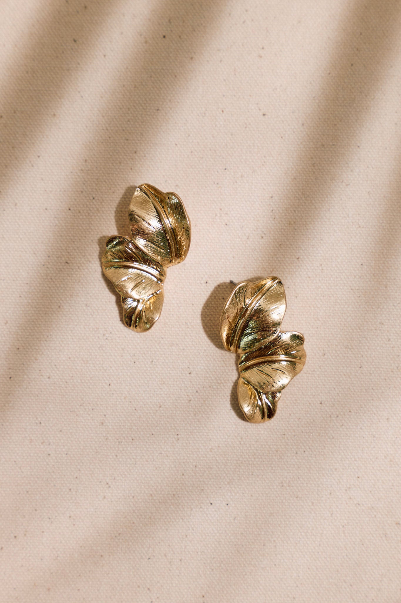 Gold palm leaf earrings displayed on a beige background emphasizing the worn gold tone and layered leaf shapes.