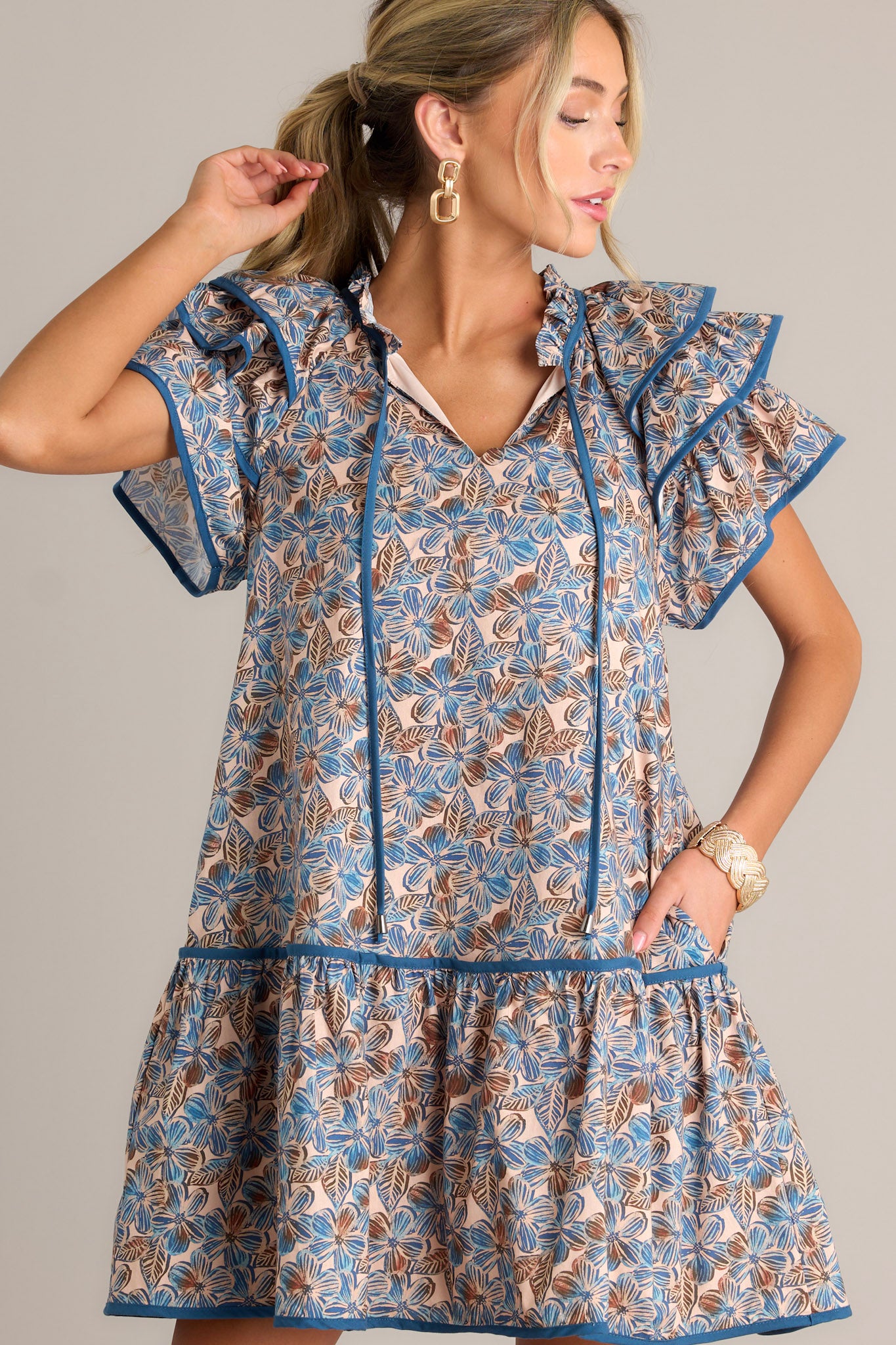 Side view of a blue floral mini dress showcasing the ruffled v-neckline with a self-tie feature, relaxed fit, functional hip pockets, single tier, and ruffled short sleeves.
