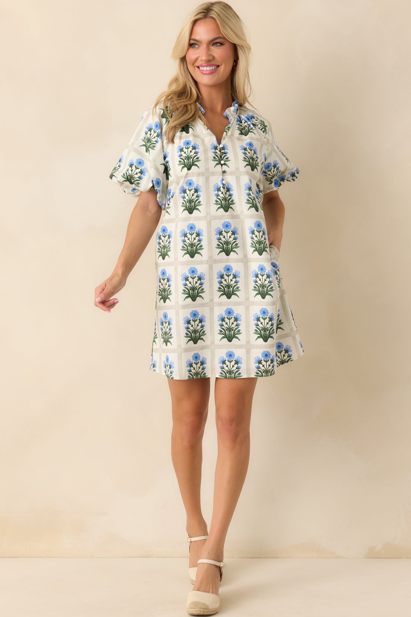 Photo of model walking in a mini dress that features a geometric grid print, blue floral motifs in each square, ruffle trim notch v-neckline, functional button front bust, short puff sleeves, elastic cuffs, ivory lining, and functional pockets.