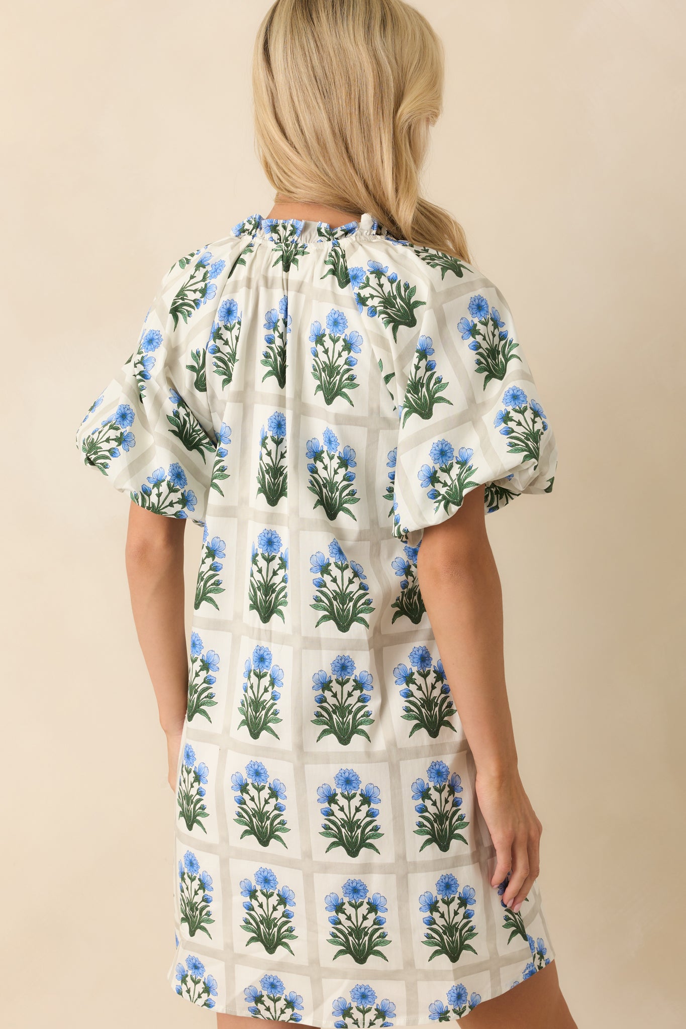 Photo of back of model wearing a mini dress that features a geometric grid print, blue floral motifs in each square, ruffle trim notch v-neckline, functional button front bust, short puff sleeves, elastic cuffs, ivory lining, and functional pockets.