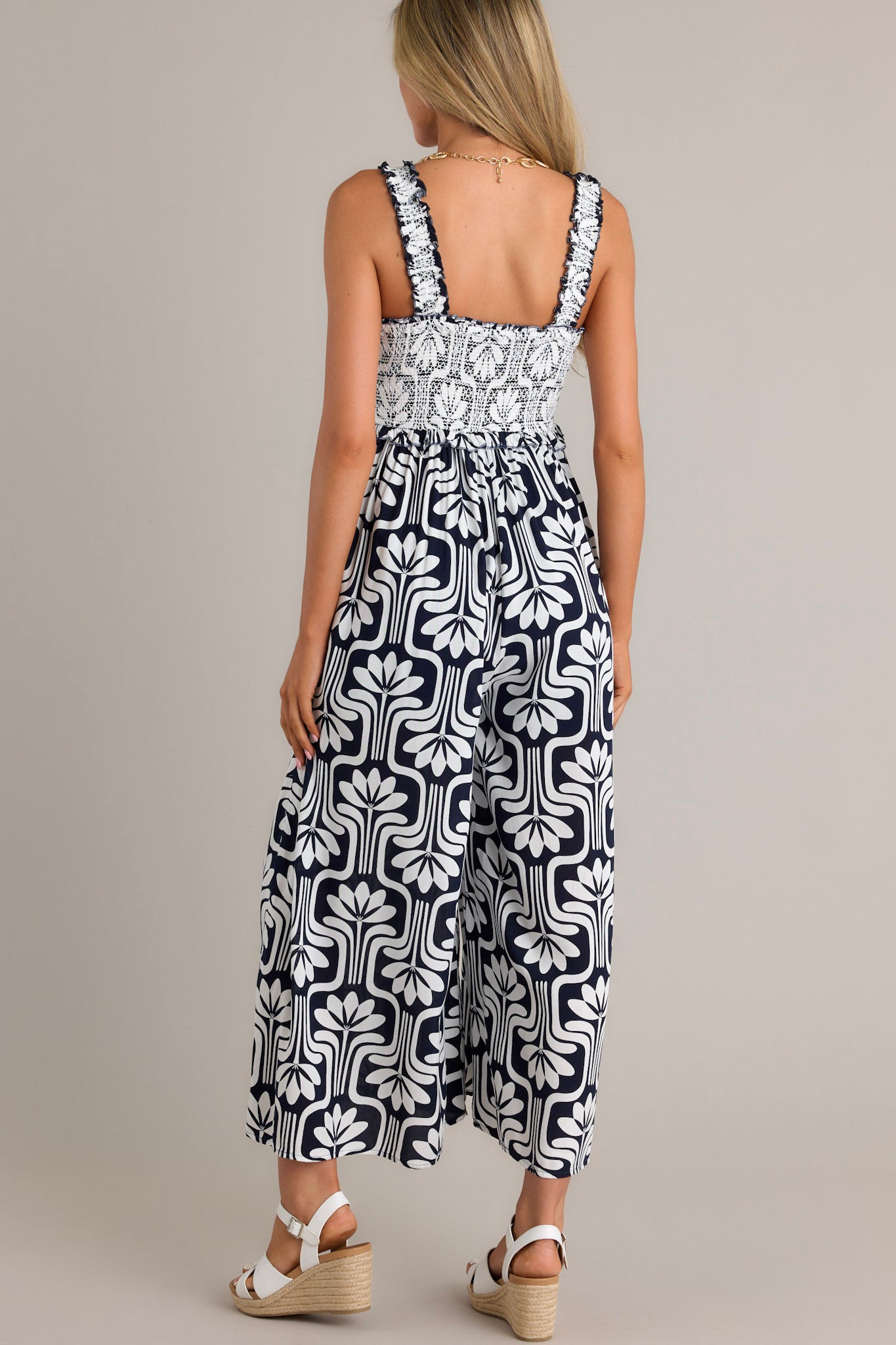 Back view of a black jumpsuit highlighting the elastic ruffle straps, fully smocked bust, and slightly cropped hemline.