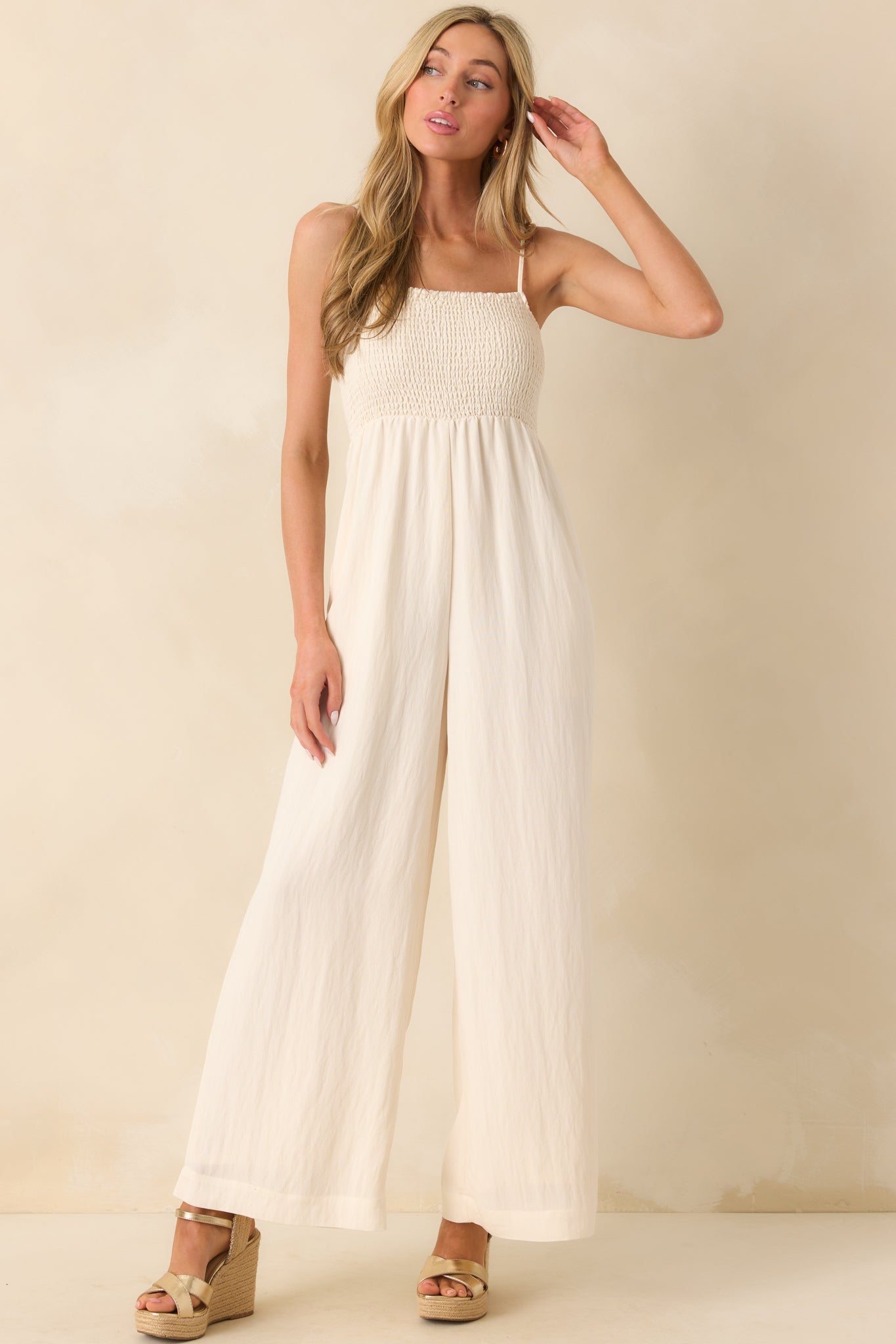 Photo of model wearing a ivory jumpsuit that features a fully smocked bust, square neckline, adjustable spaghetti straps, lightweight rayon blend fabric, wide ankle length legs, and is holding a seashell handbag.