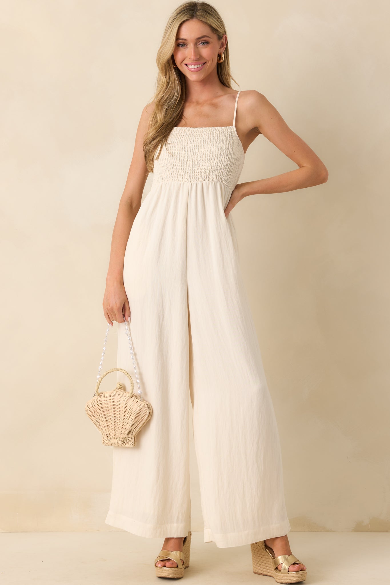 Photo of model wearing a ivory jumpsuit that features a fully smocked bust, square neckline, adjustable spaghetti straps, lightweight rayon blend fabric, wide ankle length legs, and is holding a seashell handbag.