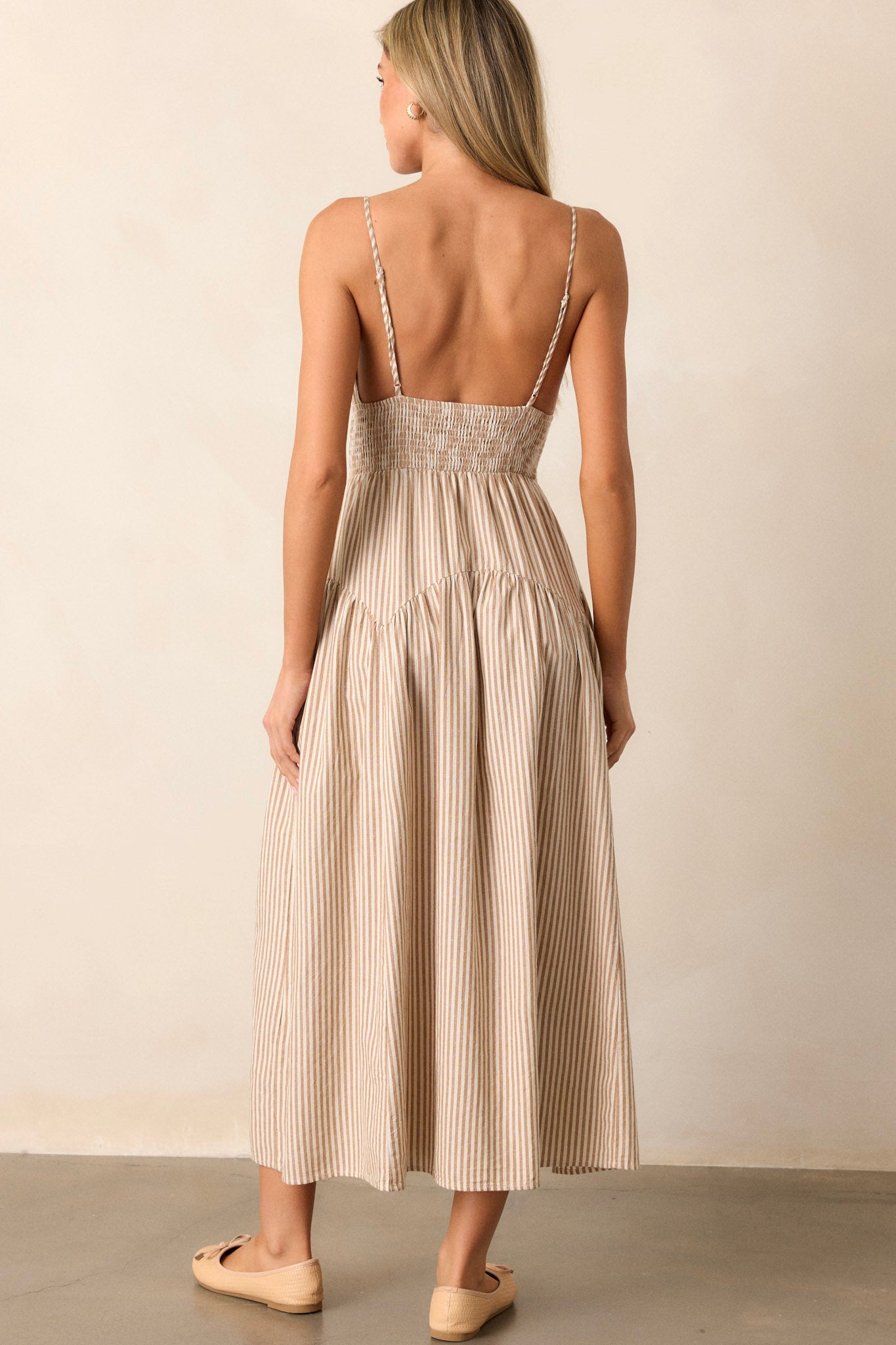 Back view of this tan midi dress that features a deep v-neckline, a smocked waistband, functional hip pockets, a stripe print, and adjustable spaghetti straps.