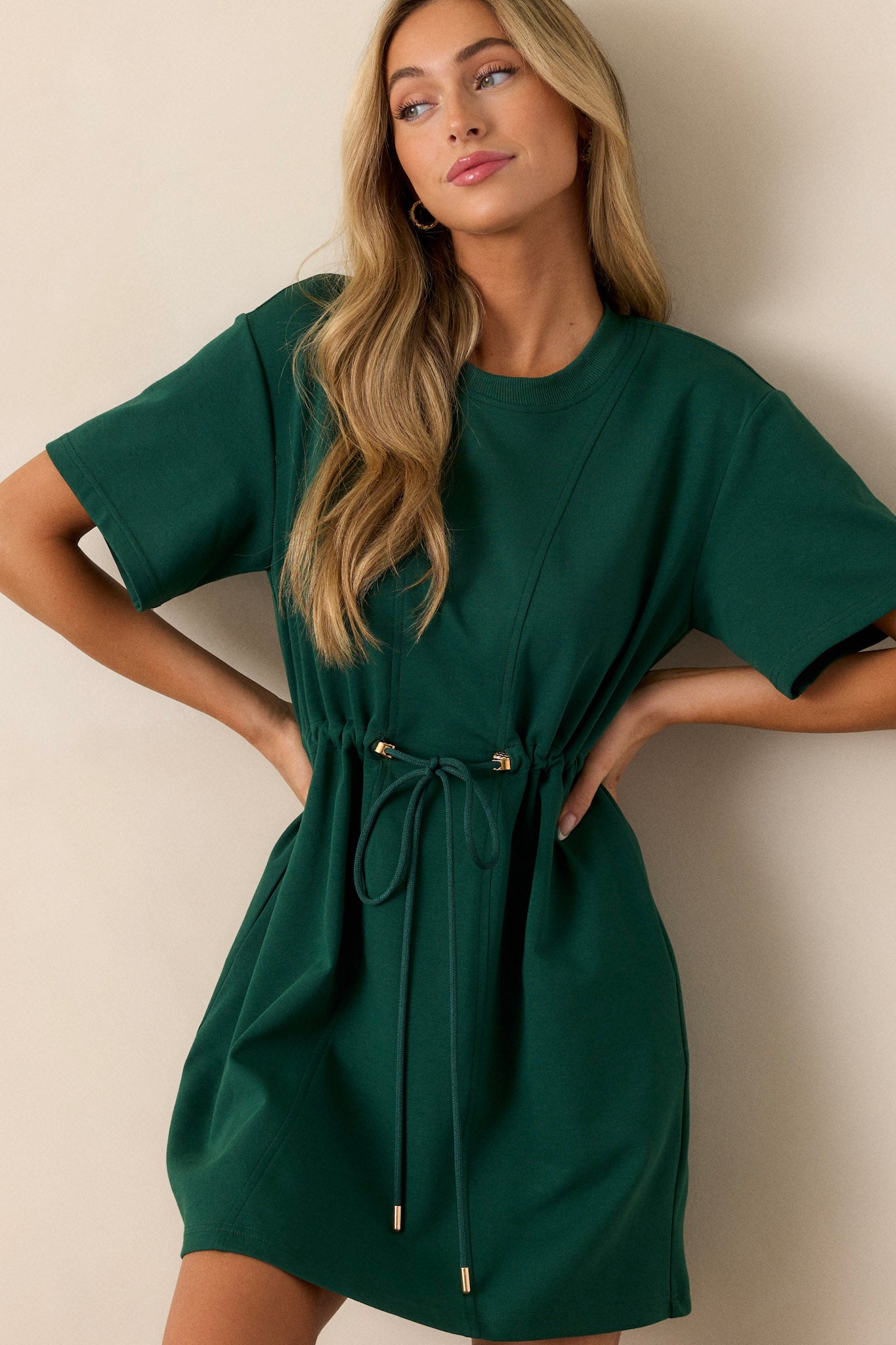 Front view of a hunter green mini dress featuring a crew neckline, a drawstring waist with gold heart shaped charms on the strings, gold aglets, two functional hip pockets, and short sleeves.