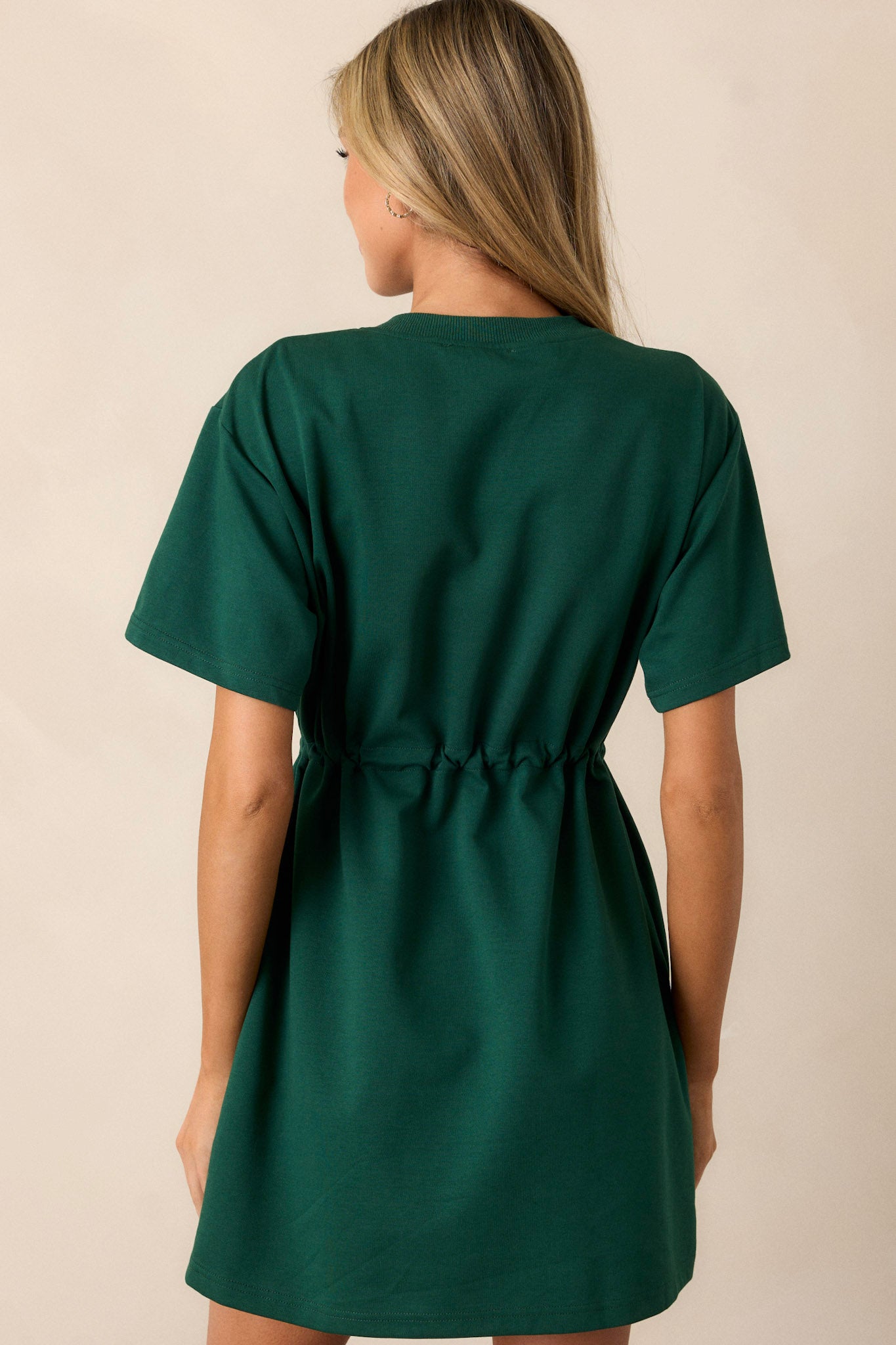 Back view of a hunter green mini dress featuring a crew neckline, a drawstring waist with gold heart shaped charms on the strings, gold aglets, two functional hip pockets, and short sleeves.
