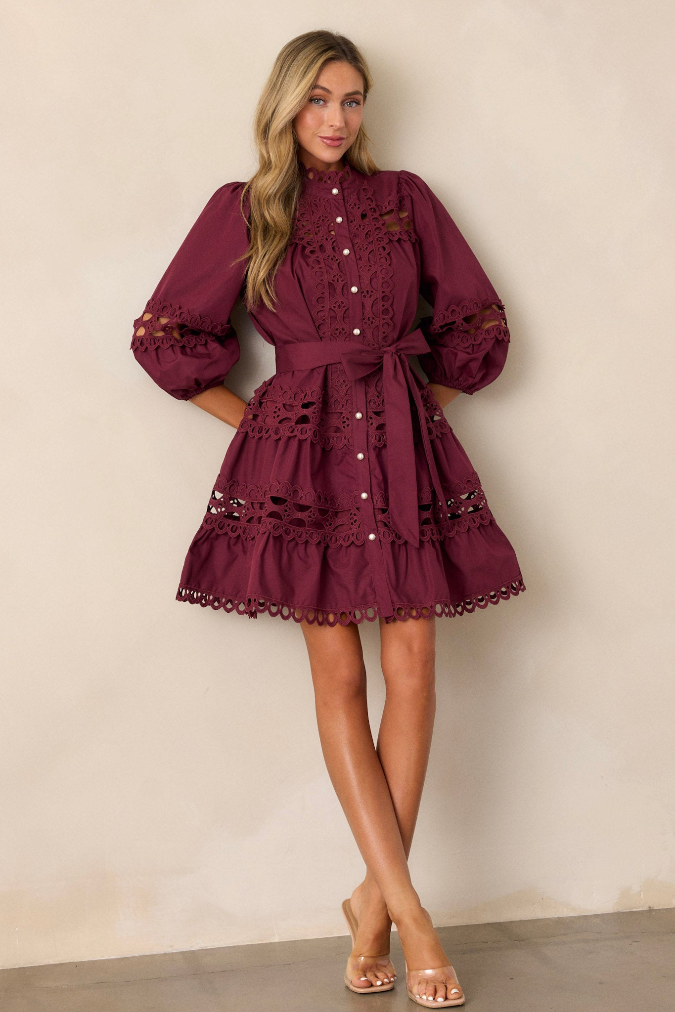 Angled view of this plum mini dress featuring a high neckline, a functional button front with faux pearl buttons, an optional self-tie belt, elastic cuffed 3/4 length sleeves, and eyelet detailing throughout.