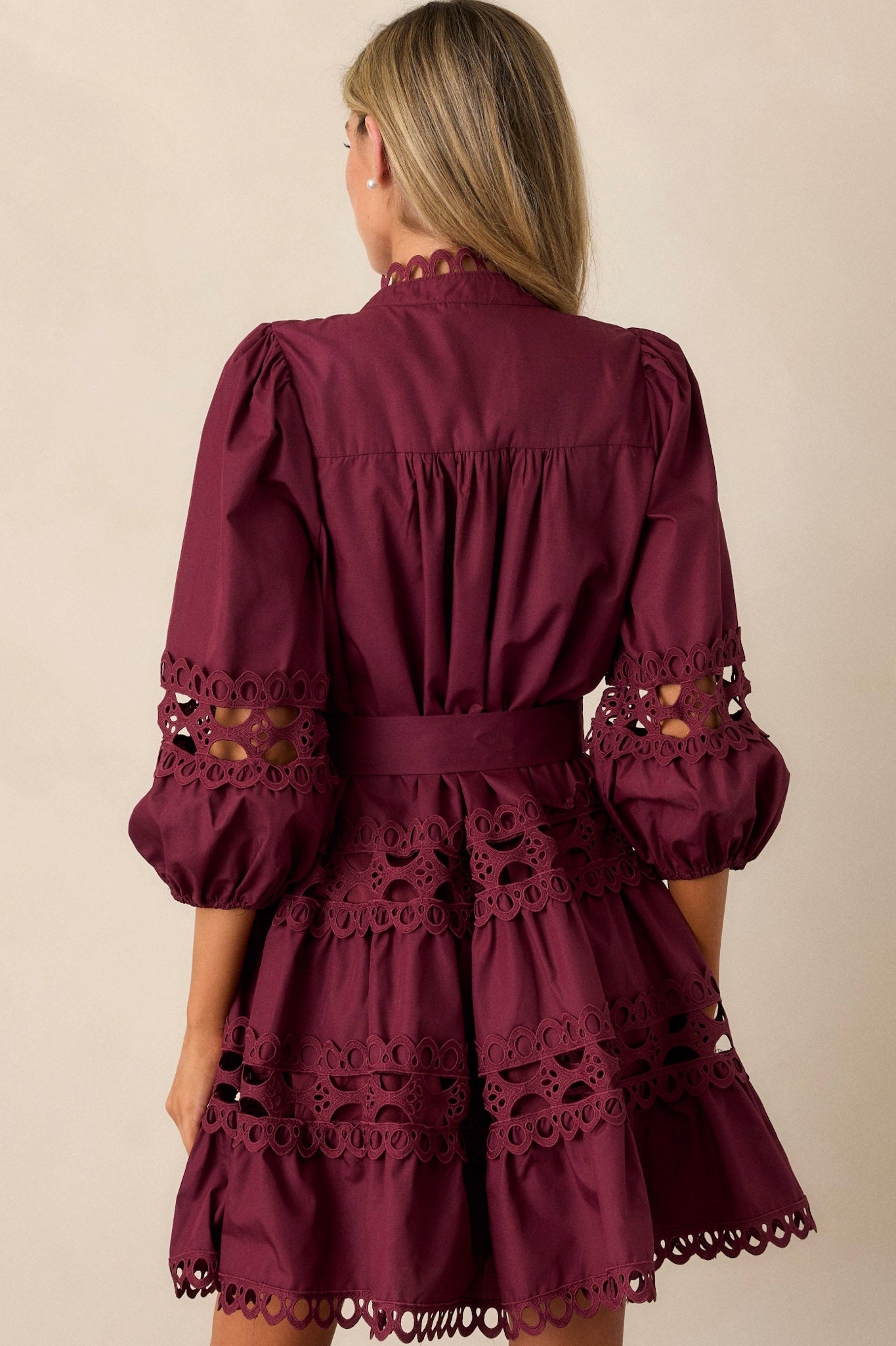 Back view of this plum mini dress featuring a high neckline, a functional button front with faux pearl buttons, an optional self-tie belt, elastic cuffed 3/4 length sleeves, and eyelet detailing throughout.