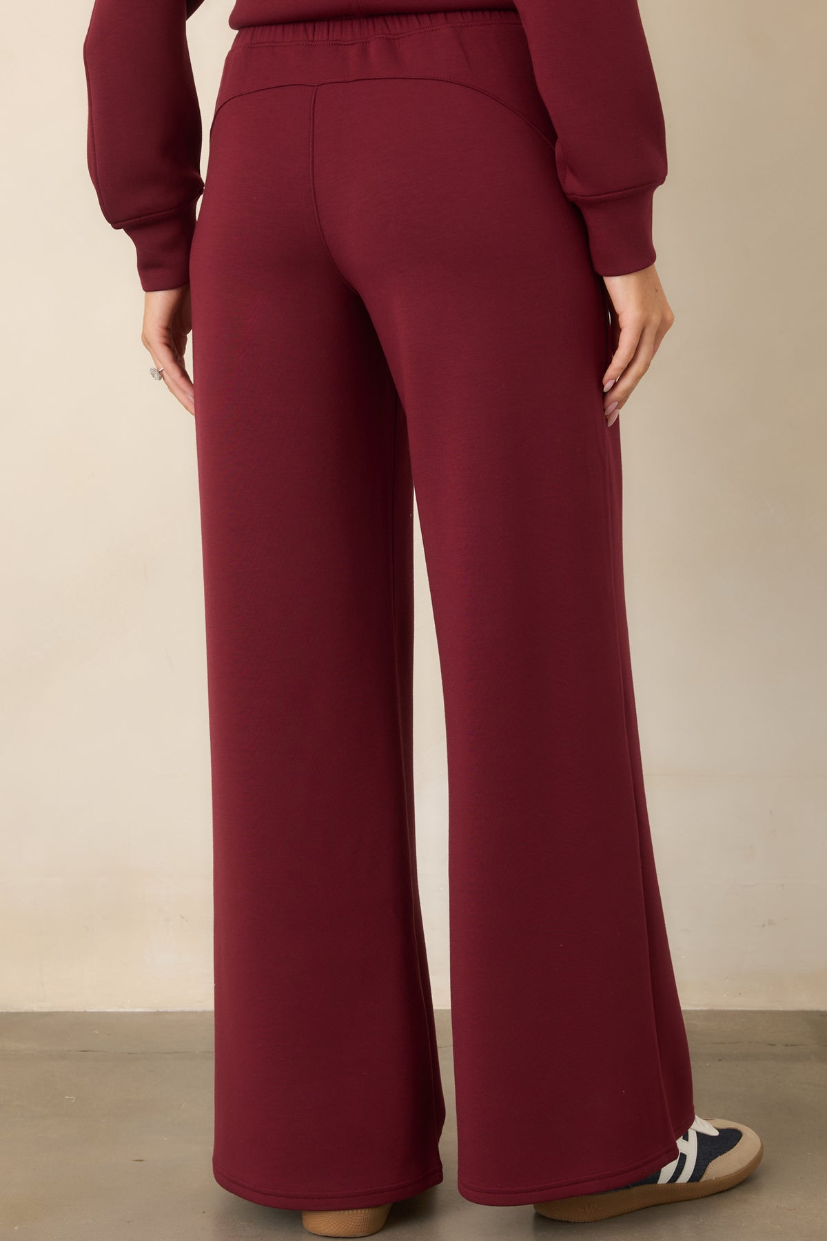 A person shown from the back wearing burgundy wide-leg sweatpants and a matching sweatshirt, standing against a plain beige wall.
