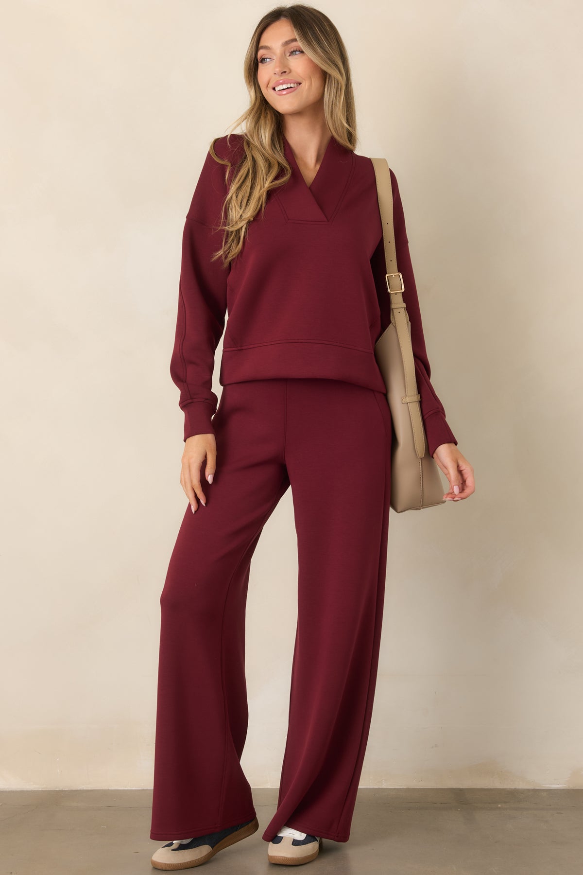 A woman modeling a matching burgundy lounge set consisting of a long-sleeve V-neck sweatshirt and wide-leg pants, styled with a beige shoulder bag and neutral sneakers against a beige studio backdrop.