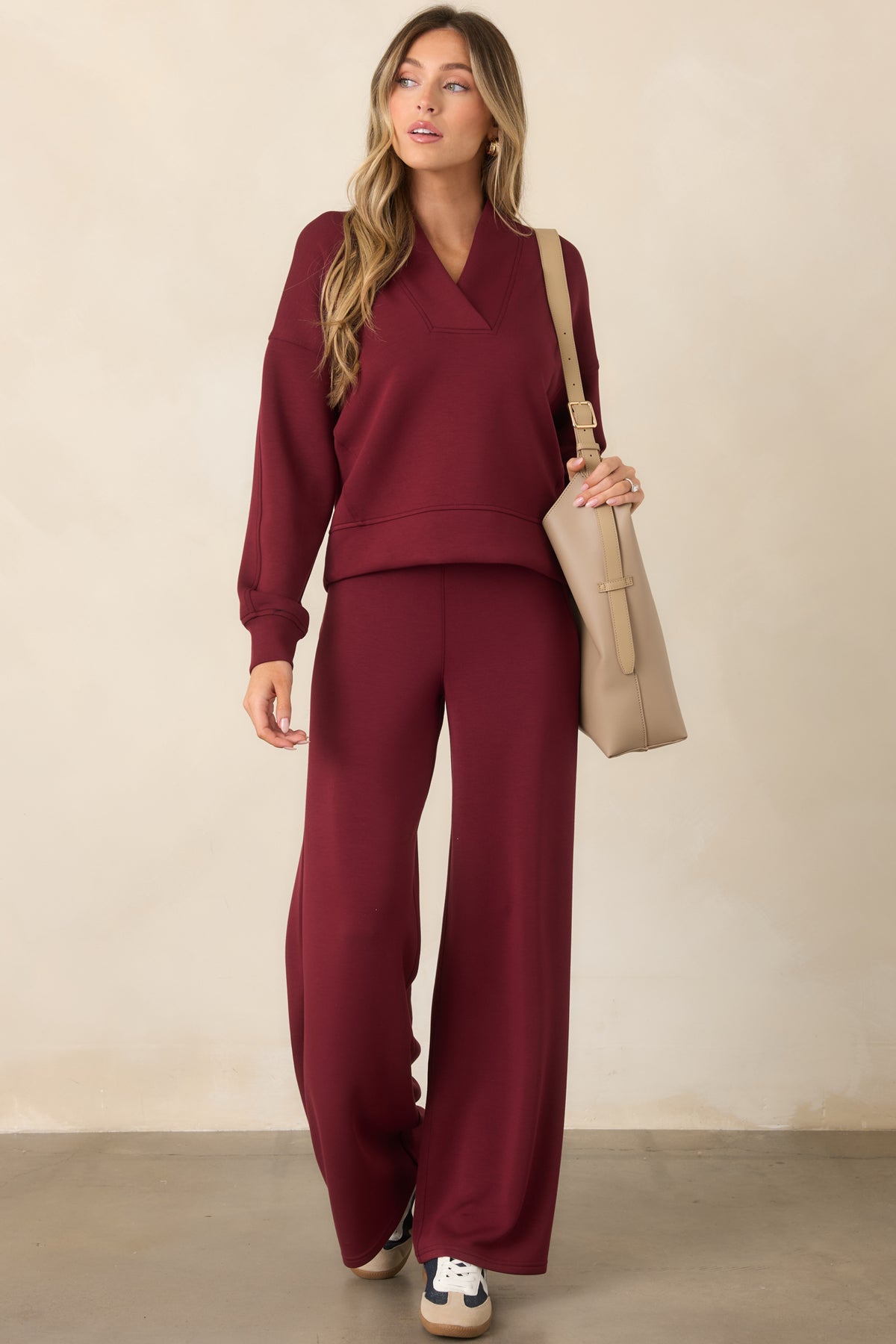 A woman modeling a matching burgundy lounge set consisting of a long-sleeve V-neck sweatshirt and wide-leg pants, styled with a beige shoulder bag and sneakers against a neutral beige studio background.
