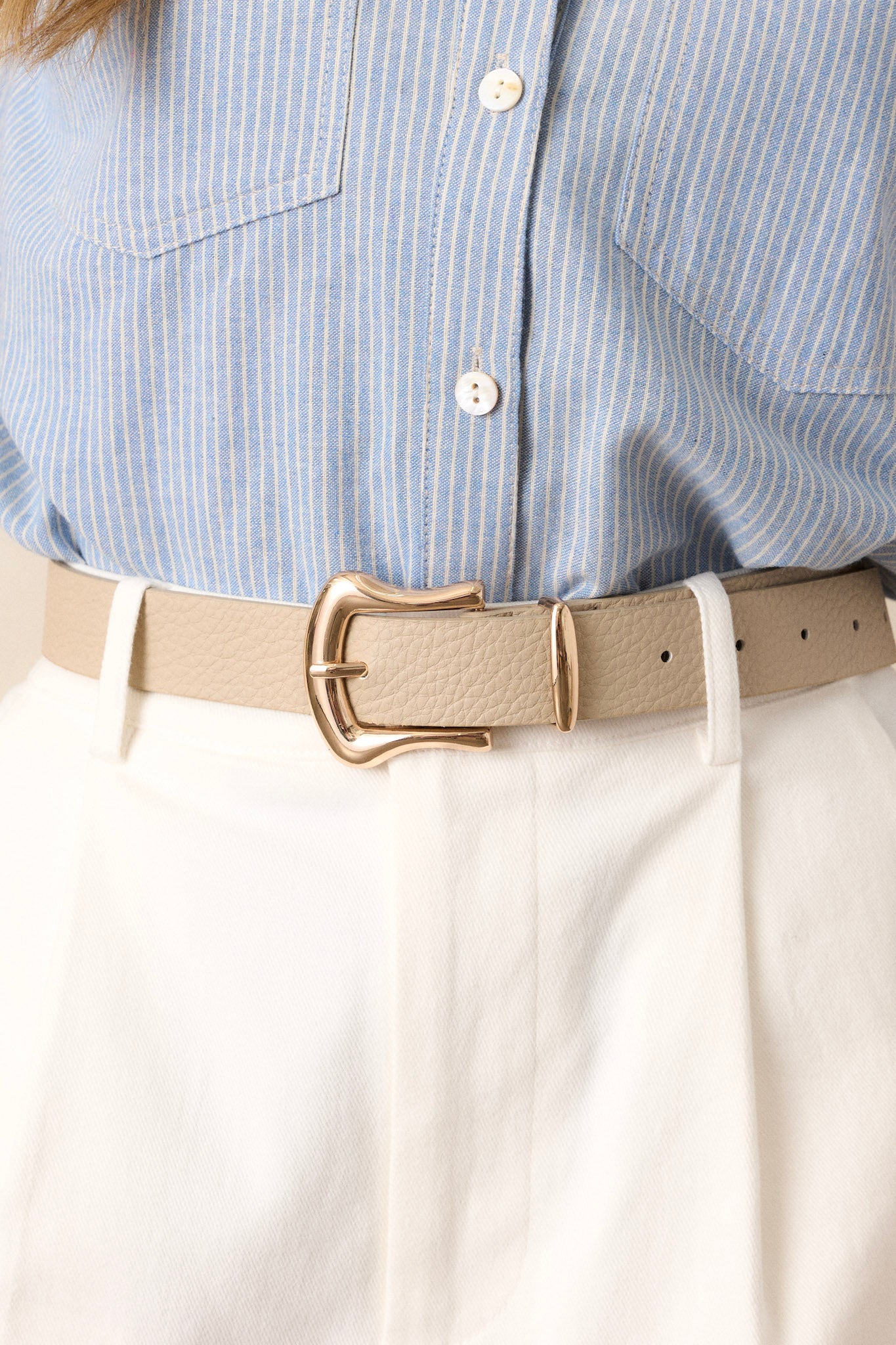 Close-up of this beige belt that features gold hardware, a horseshoe pin buckle, and a textured faux leather material