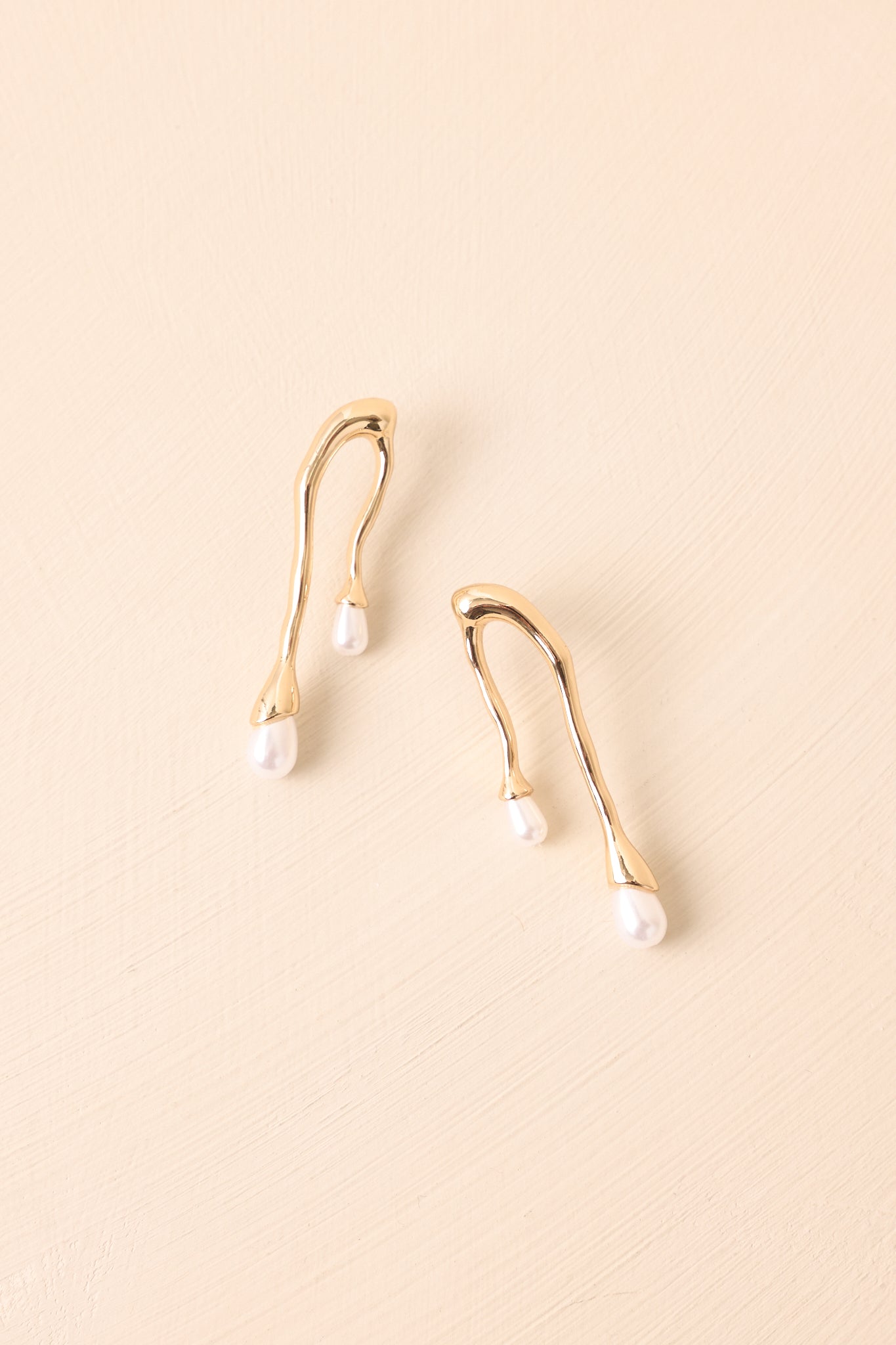 Abstract gold-tone earrings displayed on a beige background, showcasing the sculptural frame with pearl accents.
