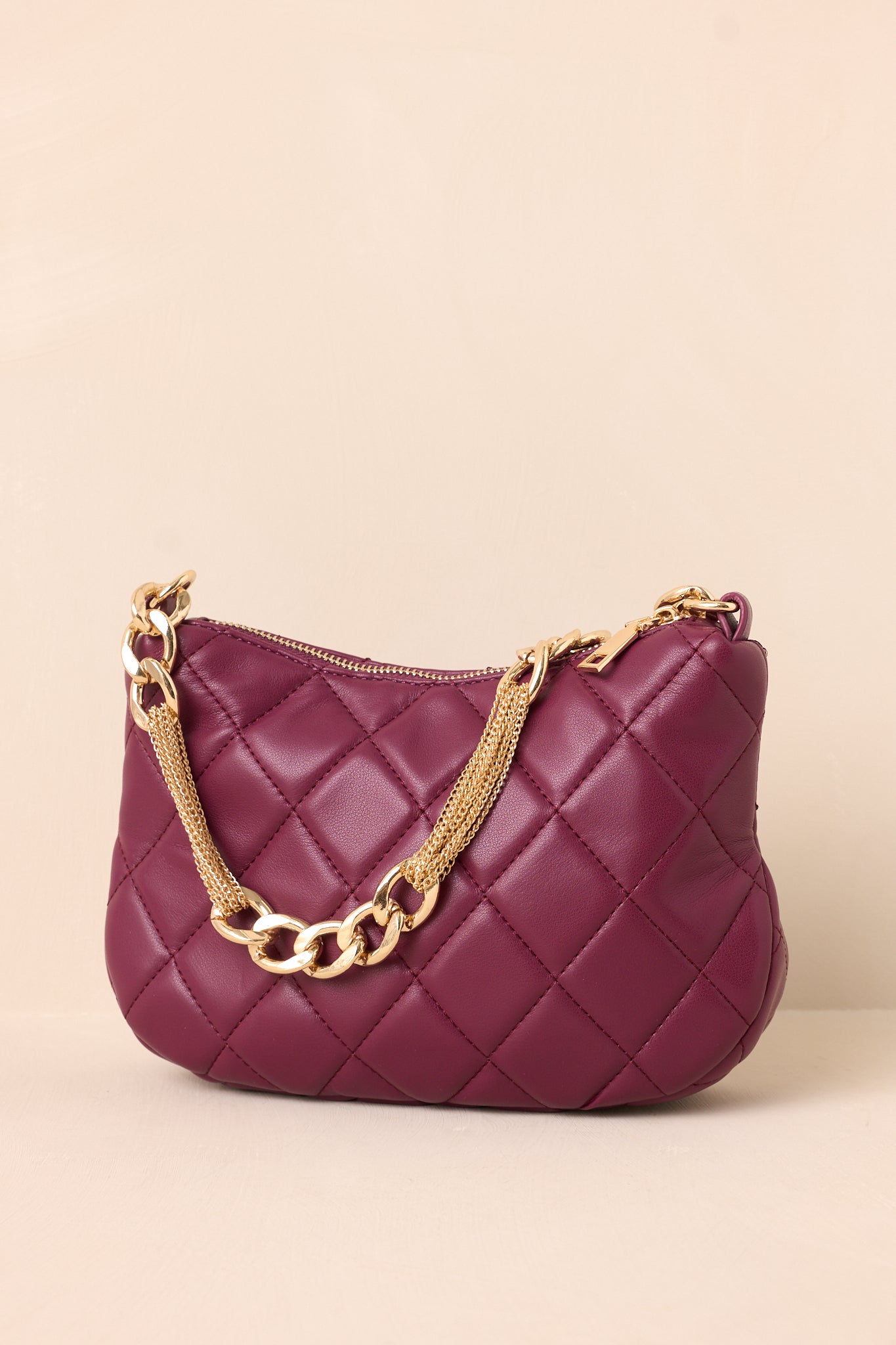 A side angle of the merlot faux leather handbag on a beige background, showing the quilted pattern and the structured silhouette with the thin gold chain shoulder strap resting nearby.