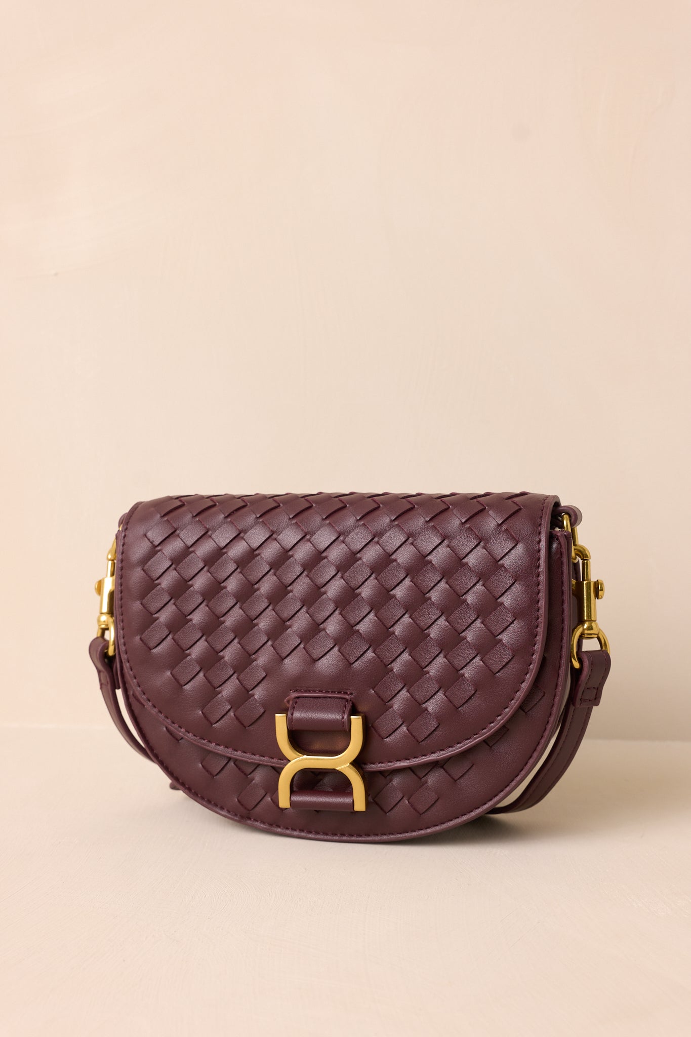 The burgundy handbag displayed upright against a beige background, showcasing its fold-over design, gold buckle snap closure, and woven faux leather material.