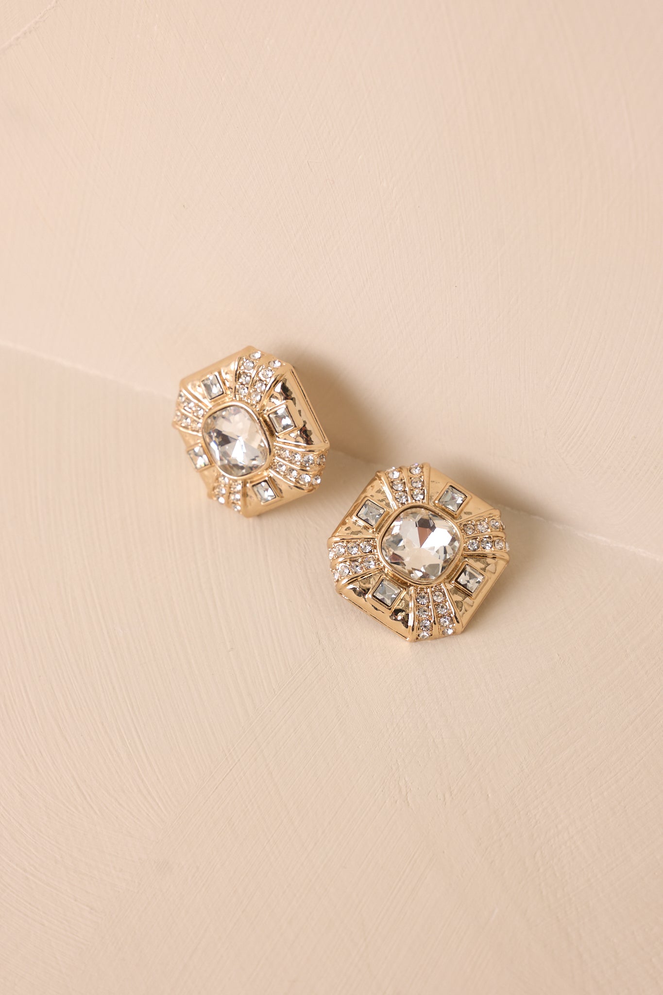 A slightly different angle of the earrings on a beige backdrop, emphasizing the large central square rhinestone and surrounding accents.