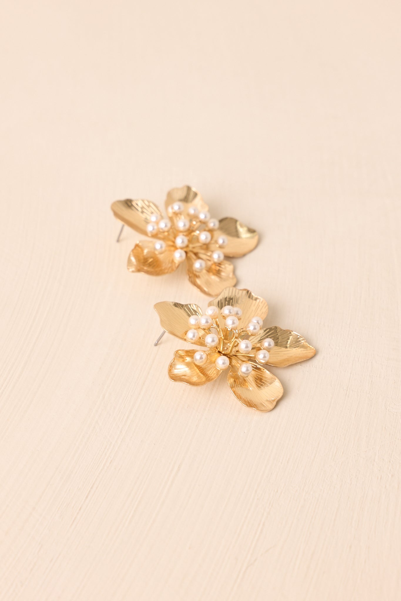 An angled shot of the floral gold earrings featuring five textured petals and a pearl cluster center shown against a beige background.