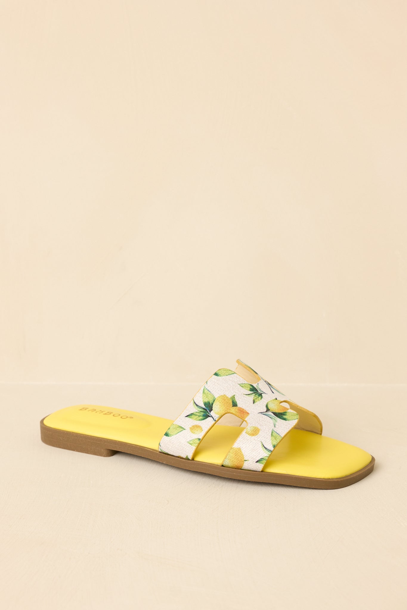 Angled view of fruit print sandals on neutral beige backdrop highlighting colorful pattern and slip-on style