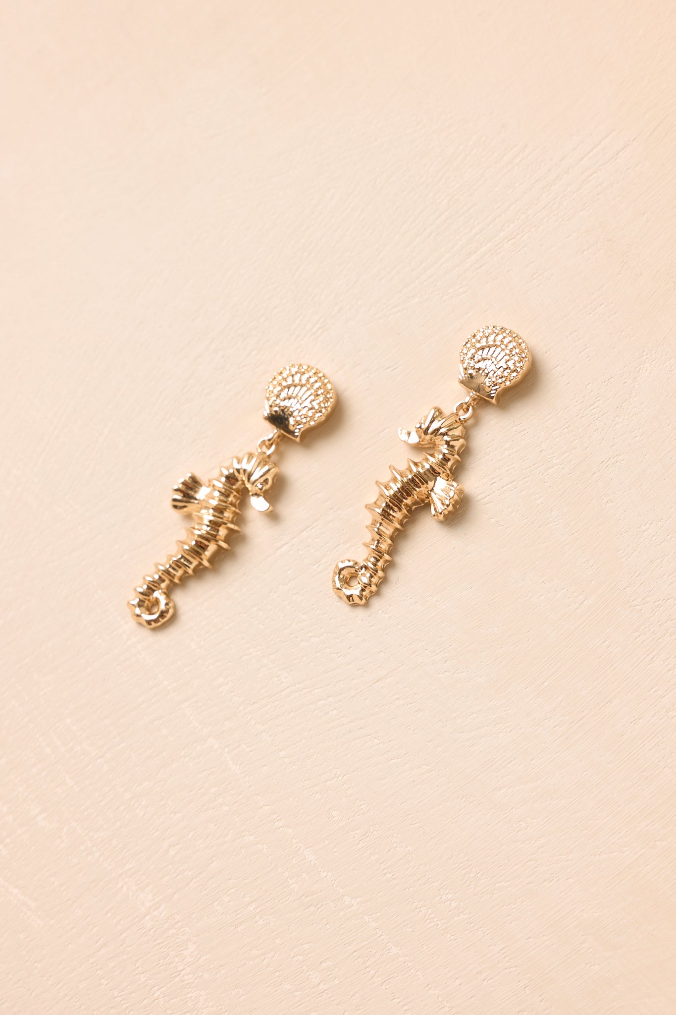 Angled view of gold earrings on a beige background highlighting the detailed seahorse charm and sun-kissed metallic sheen.