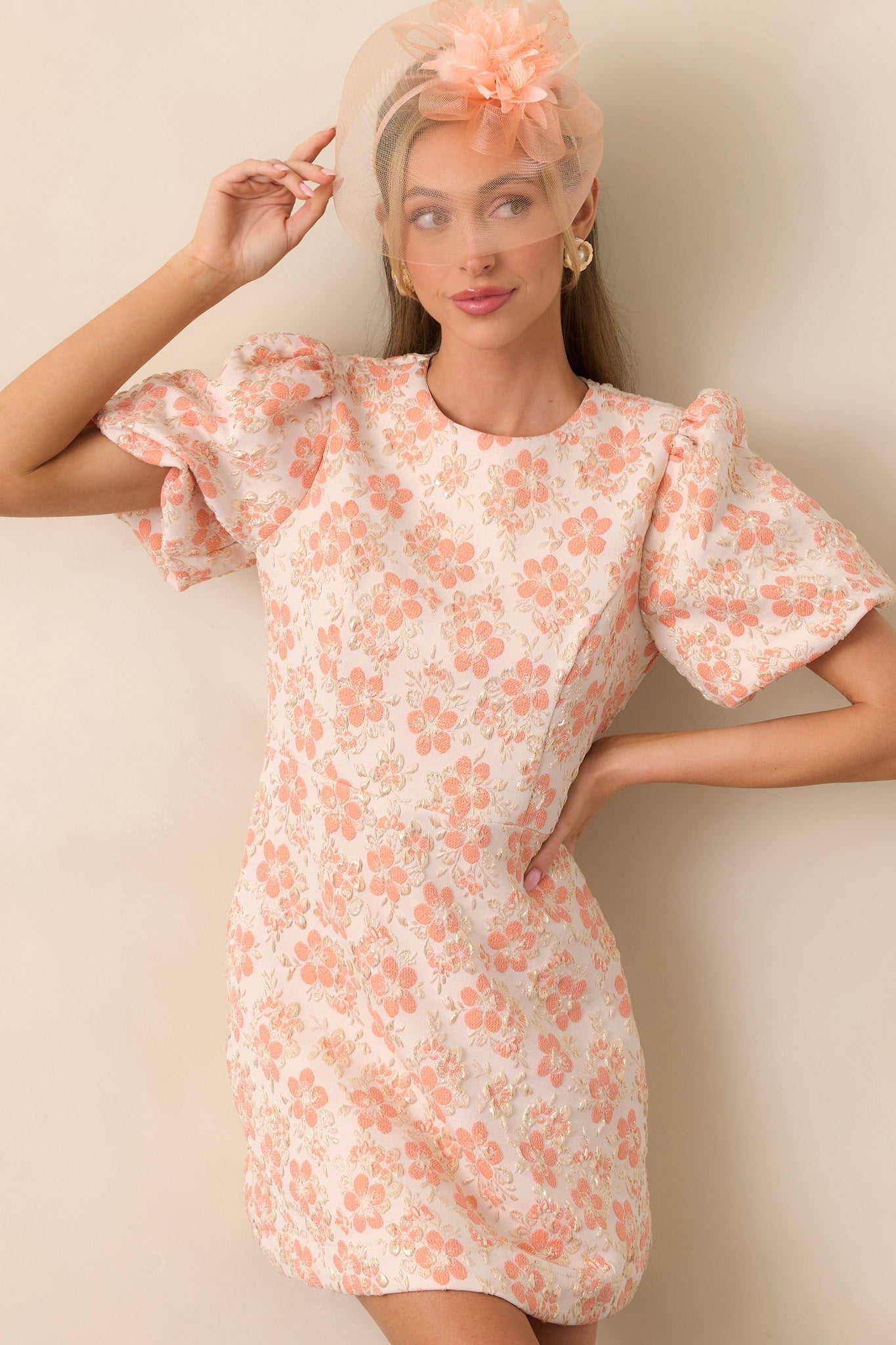 An apricot mini dress with voluminous puff sleeves, shimmering gold detailing, and a round neckline. The lightweight fabric moves effortlessly, enhancing the elegant silhouette.