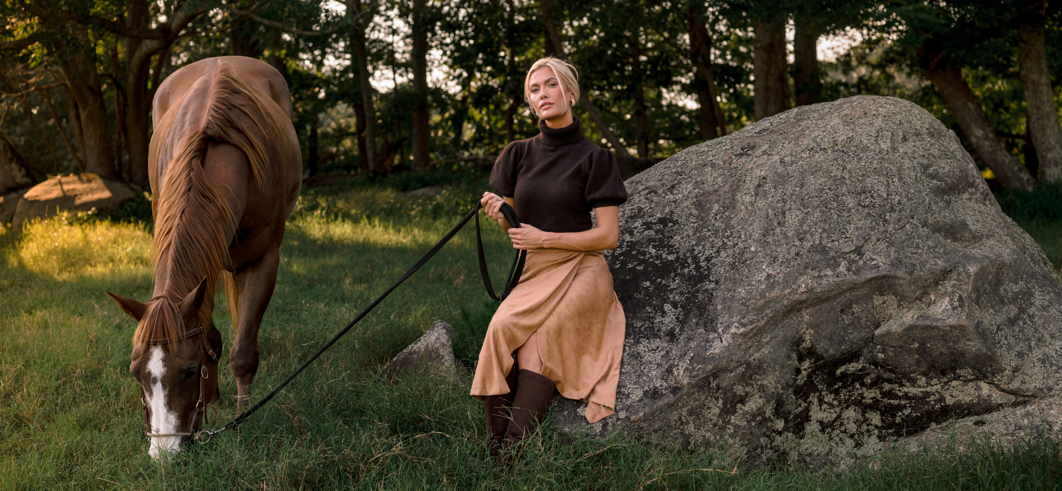 A woman in a brown turtleneck and beige skirt sits beside a large rock in a wooded outdoor setting, holding a lead attached to a brown horse grazing nearby.