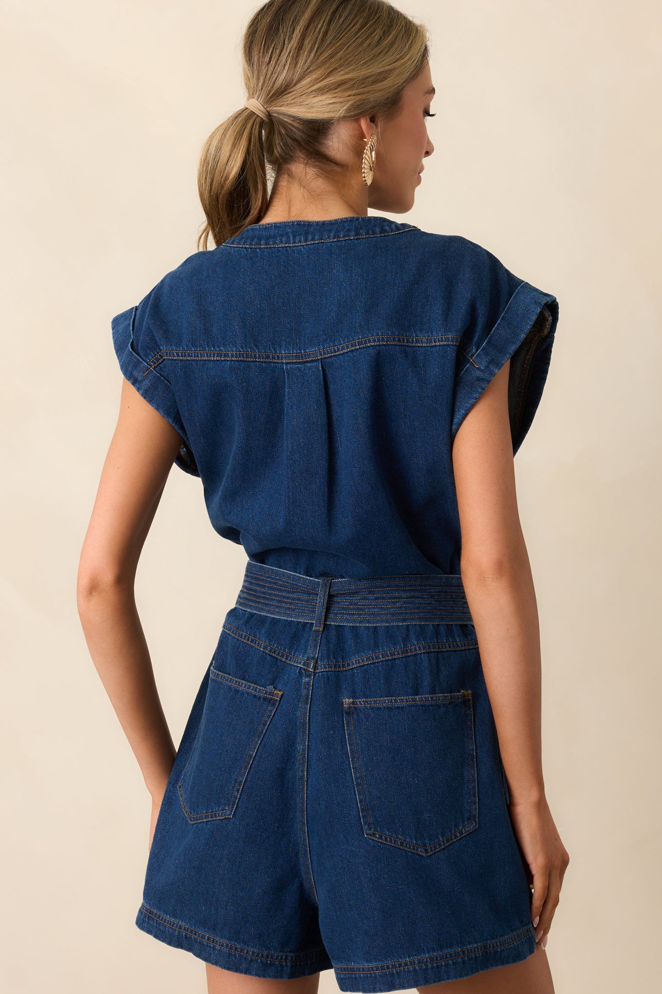 The back view of the dark wash denim romper, highlighting the simple, clean lines, functional pockets, and the stylish cap sleeves, giving a polished, laid-back look.