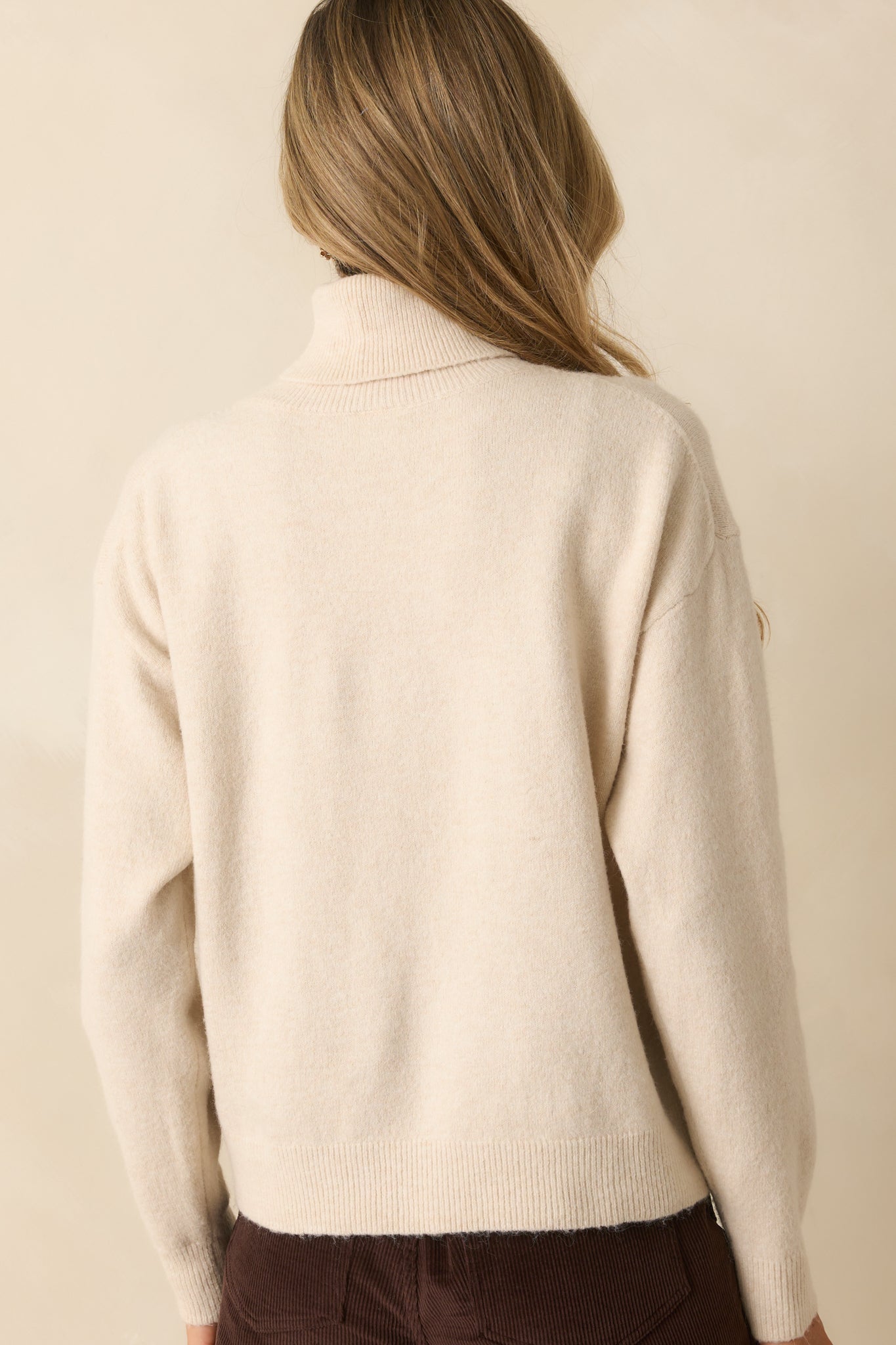 Back view of the light beige sweater showing long sleeves, fold-over turtleneck, and relaxed silhouette.