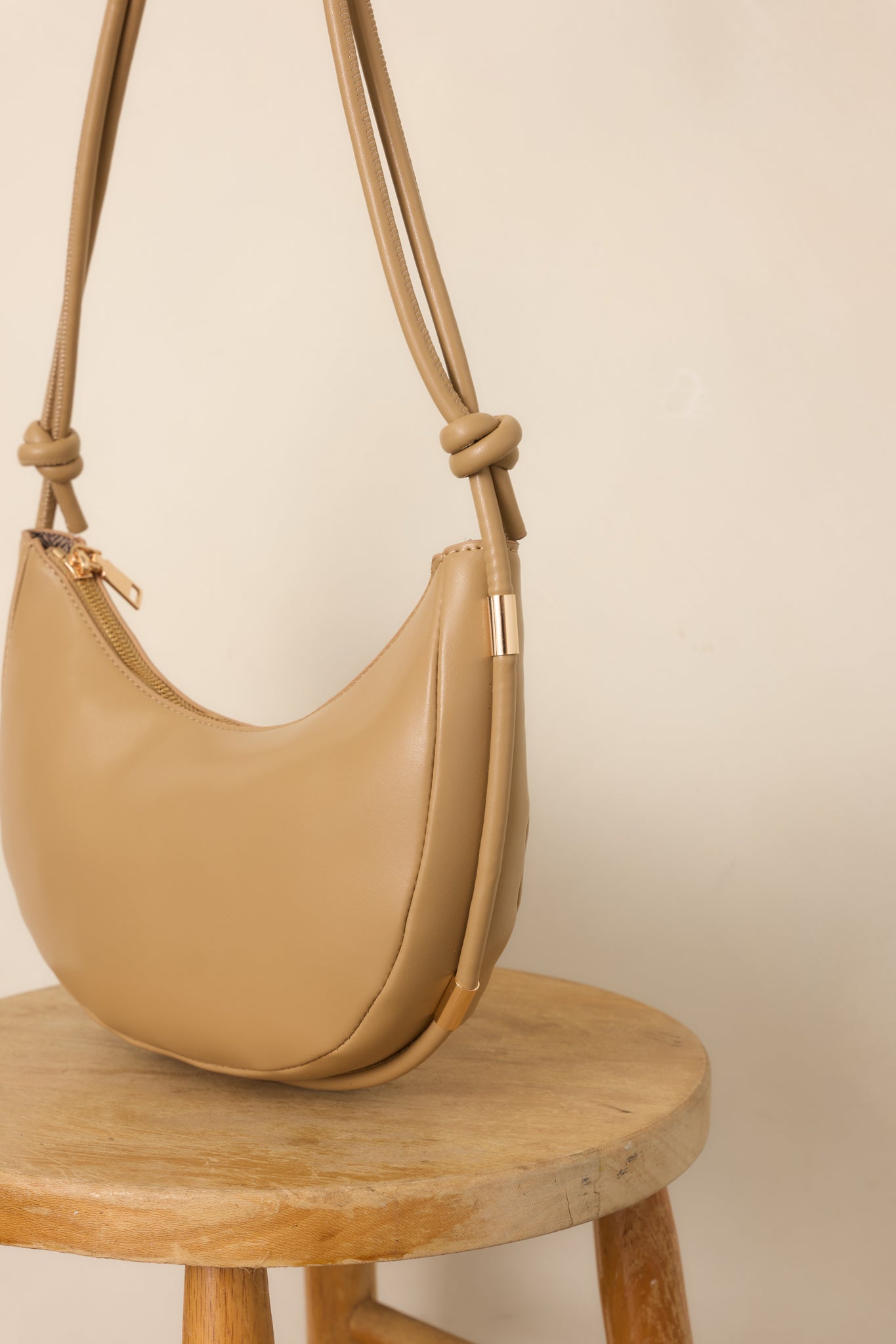Beige handbag featuring a secure top zipper closure and adjustable strap with decorative knot detailing.