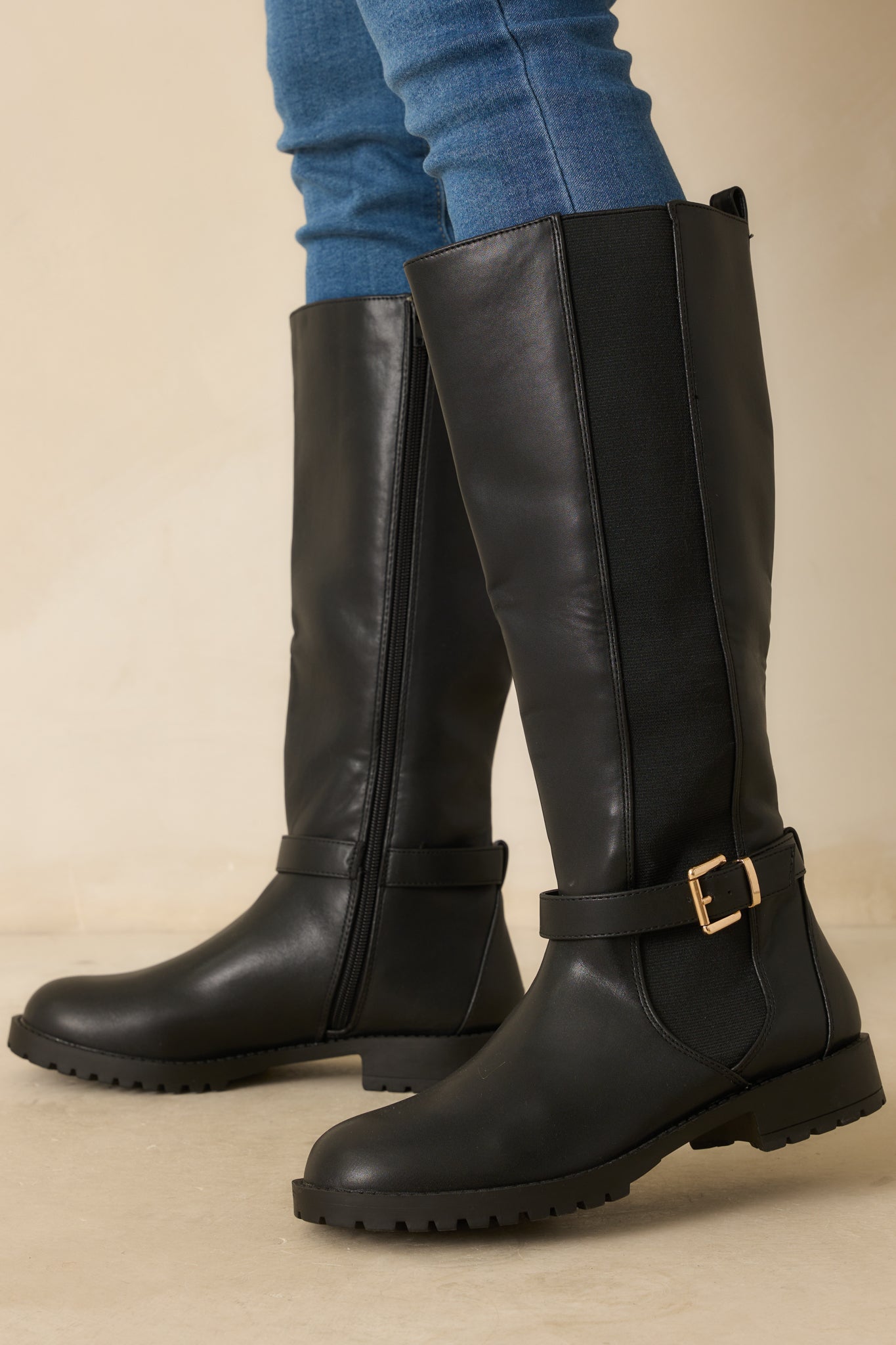 Black knee-high riding boot with smooth faux leather, round toe, and decorative gold-buckled ankle strap.